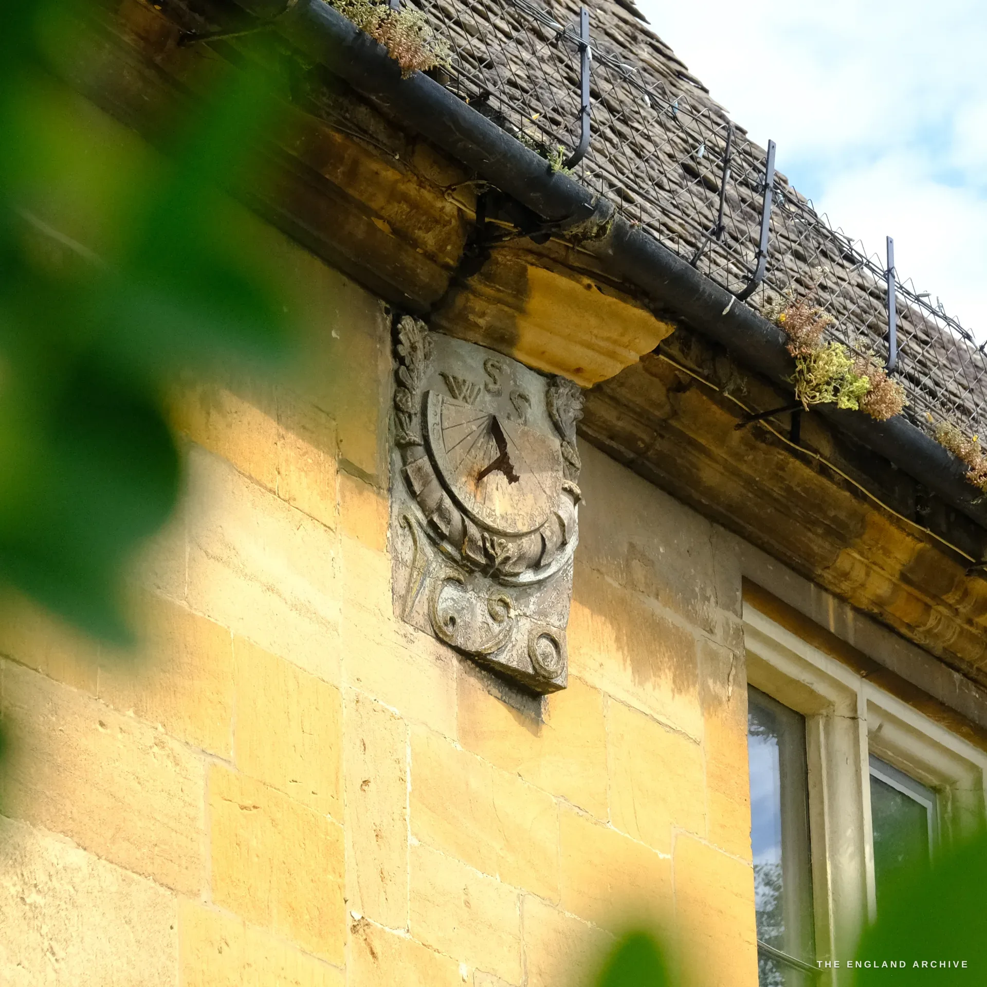A carved stone sundial set on the gable of a stone house below a stone-tiled roof, dated 1690, seen past green foliage.