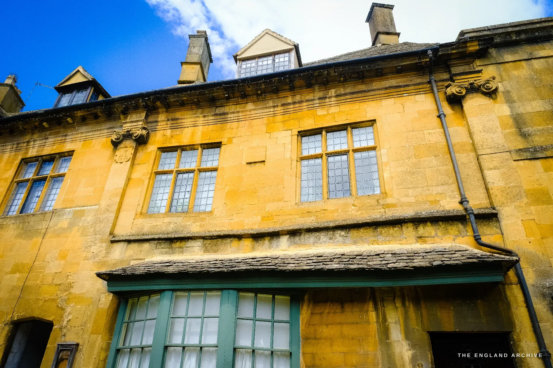 Looking up the front of a tall honey-stone house with mullioned windows, gabled dormers, carved scroll brackets and a green-painted bay window below.