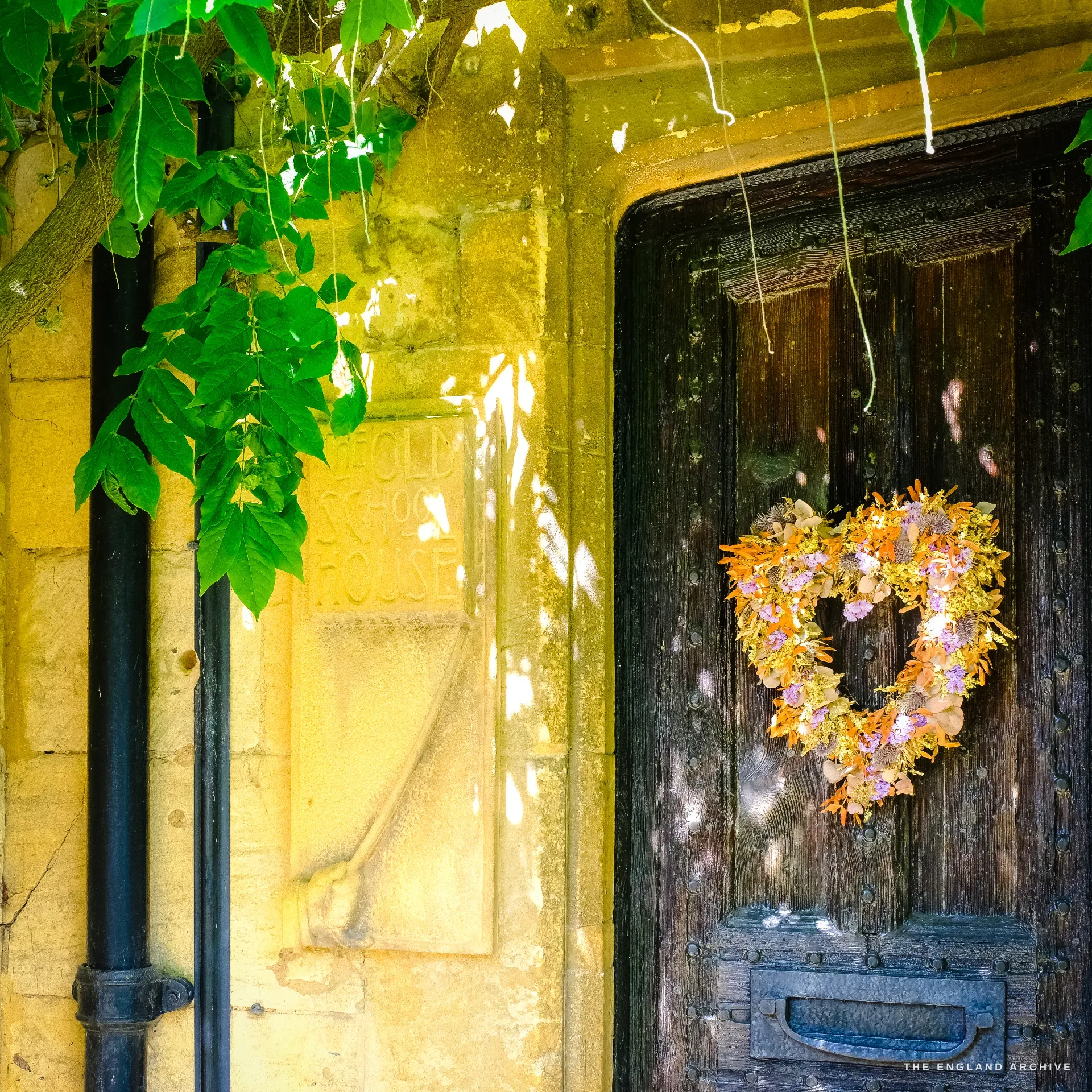 A sunlit honey-stone doorway with a dark plank door, a heart-shaped dried-flower wreath and an iron letter slot, wisteria overhanging, a worn stone tablet above reading 'The Old School House'.
