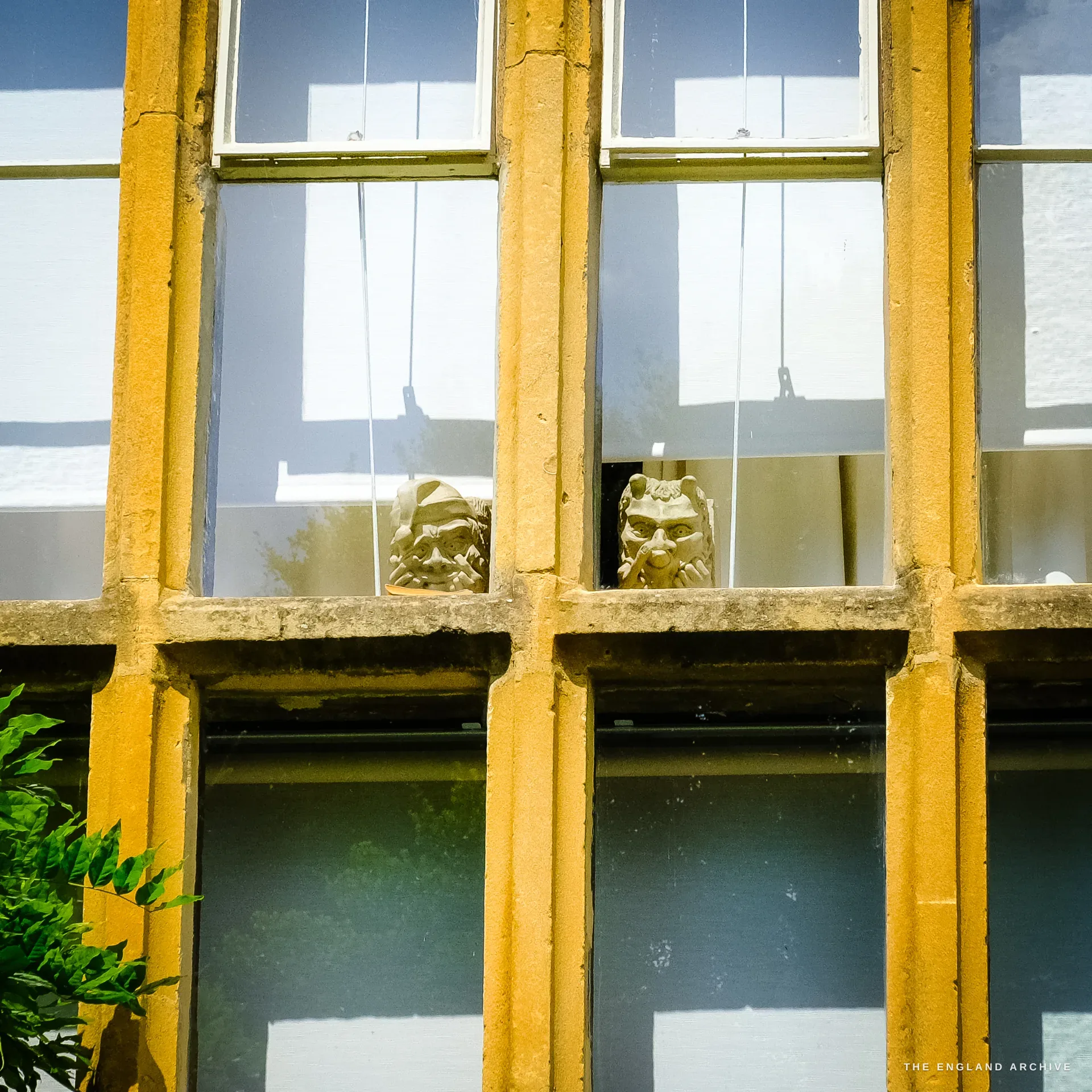 Two carved stone grotesque heads sitting on the interior sill of a tall stone-mullioned window, seen from outside through the glass.