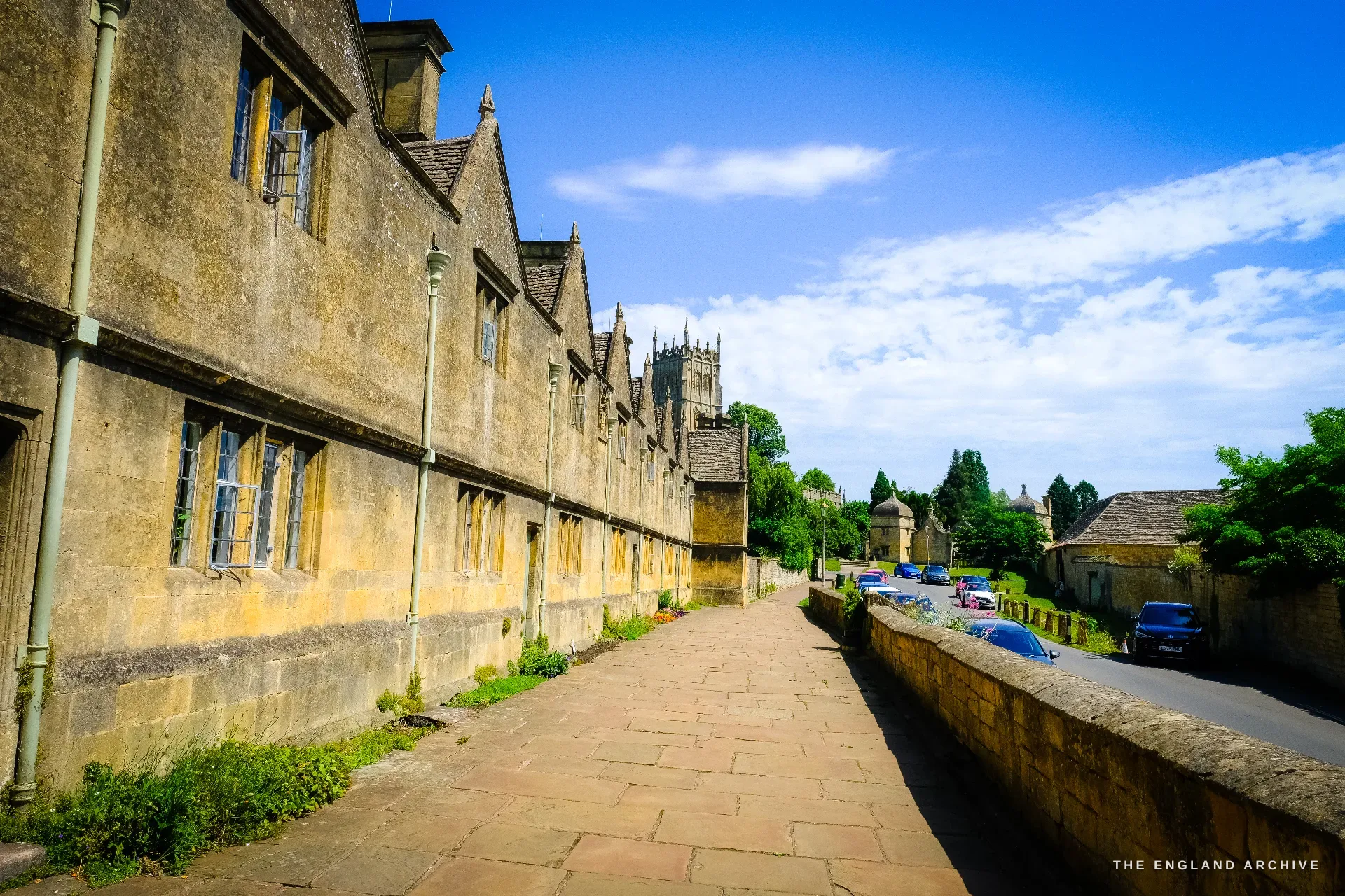 The long terrace of gabled stone almshouses on Church Street, running along a raised flagstone pavement with the tower of St James' Church beyond.