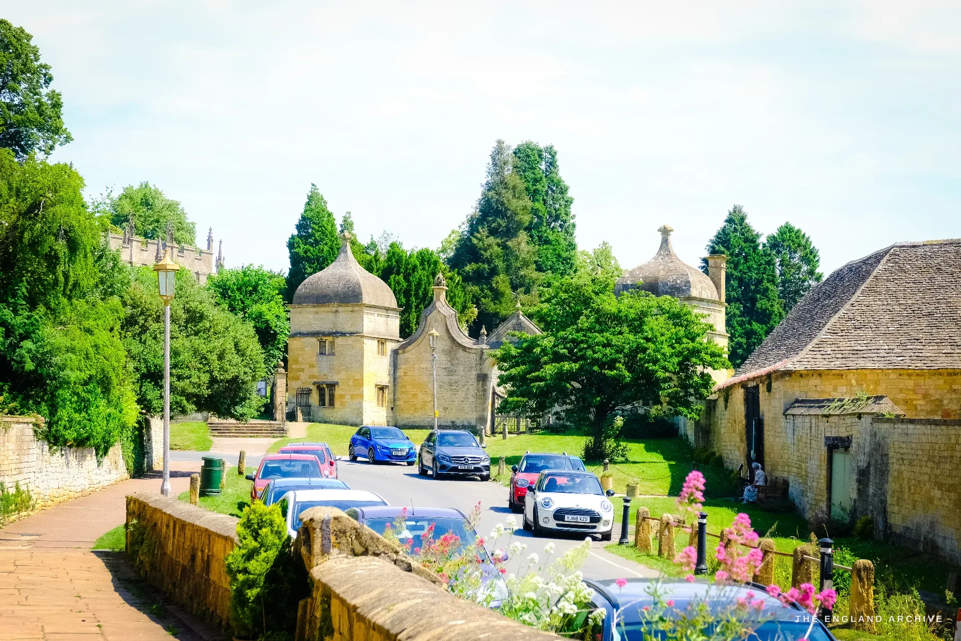 A view along Church Street toward the conical-roofed banqueting lodges of old Campden House, the church tower at left, cars parked and flowers in the foreground.