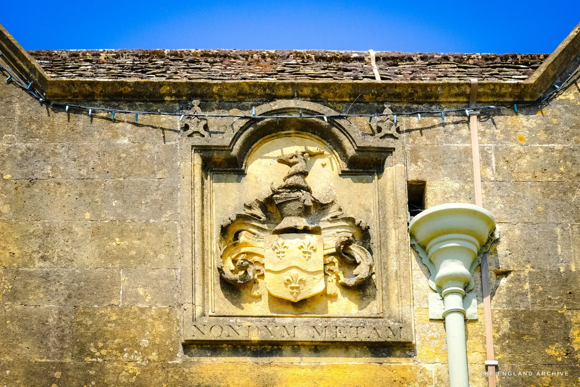 A carved heraldic panel below the eaves of a stone gable - a helm, mantling and a shield of three fleurs-de-lis - incised beneath with the Hicks motto 'Nondum metam'.