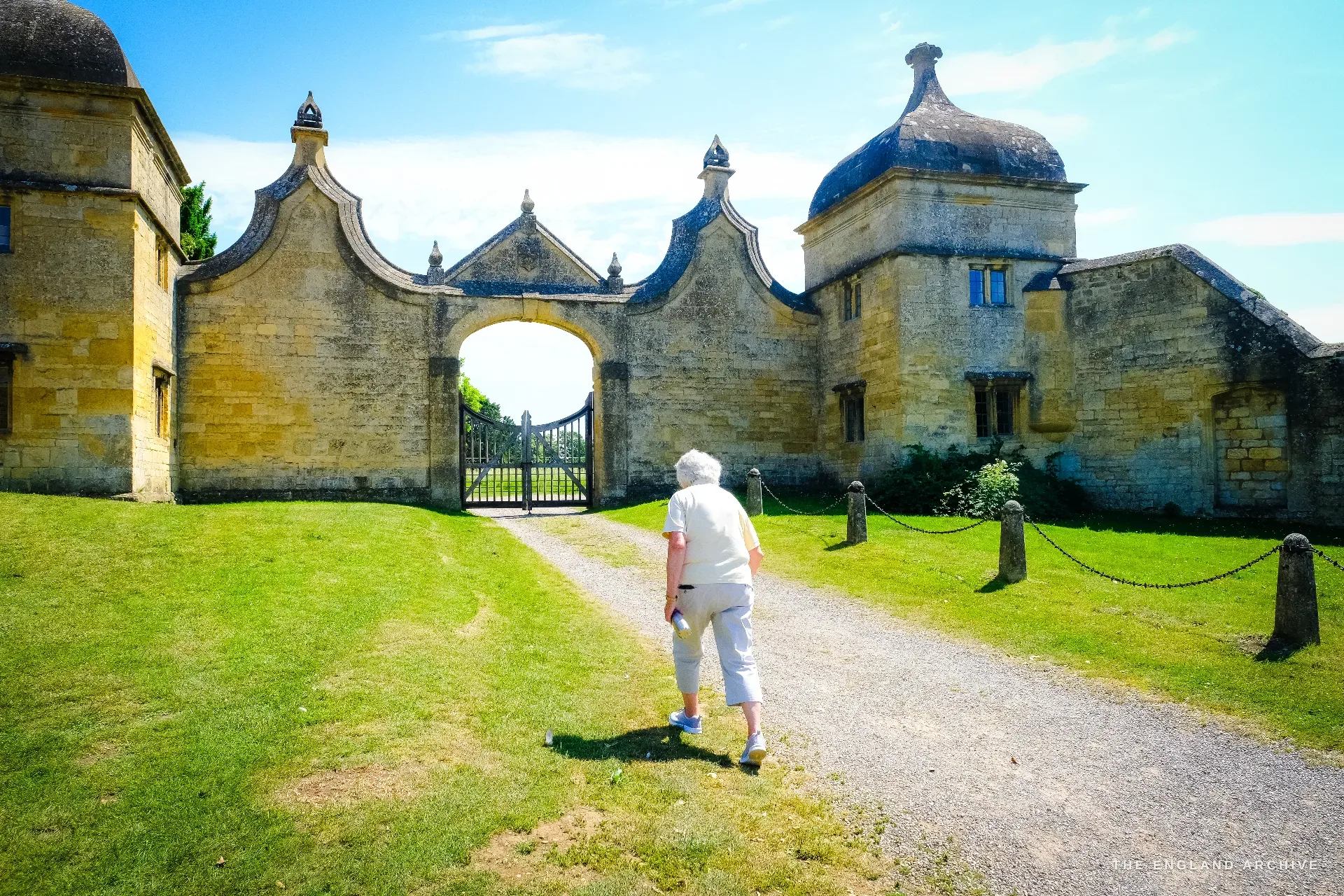 One of the ornate Jacobean gatehouse lodges of old Campden House, with ogee gables and a domed pavilion; Carol walks toward the iron gate across the lawn.