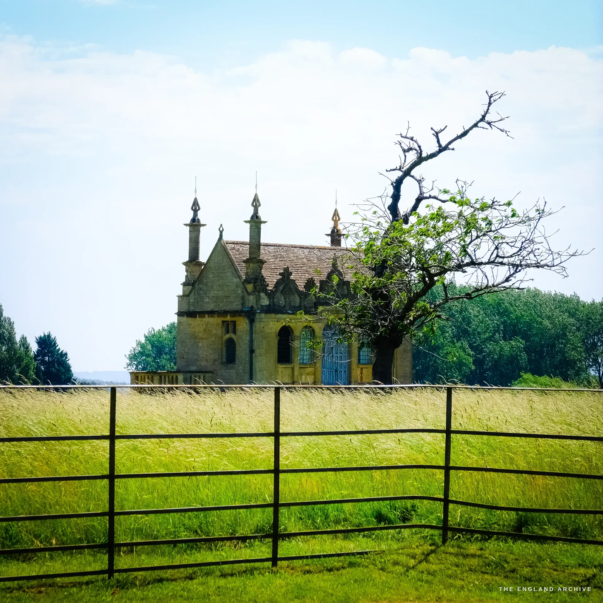 A Jacobean banqueting house of old Campden House, ornate gables and tall finial pinnacles, seen across a meadow behind a black field gate beside a gnarled tree.