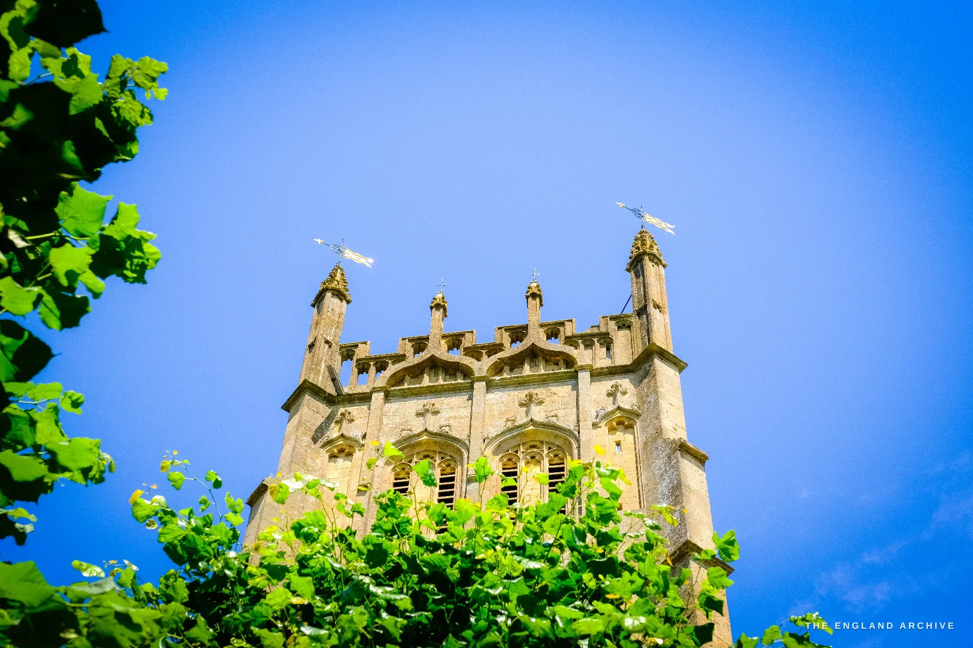 Looking up at the crocketed, pinnacled Perpendicular top of St James' Church tower against blue sky, framed by green foliage.