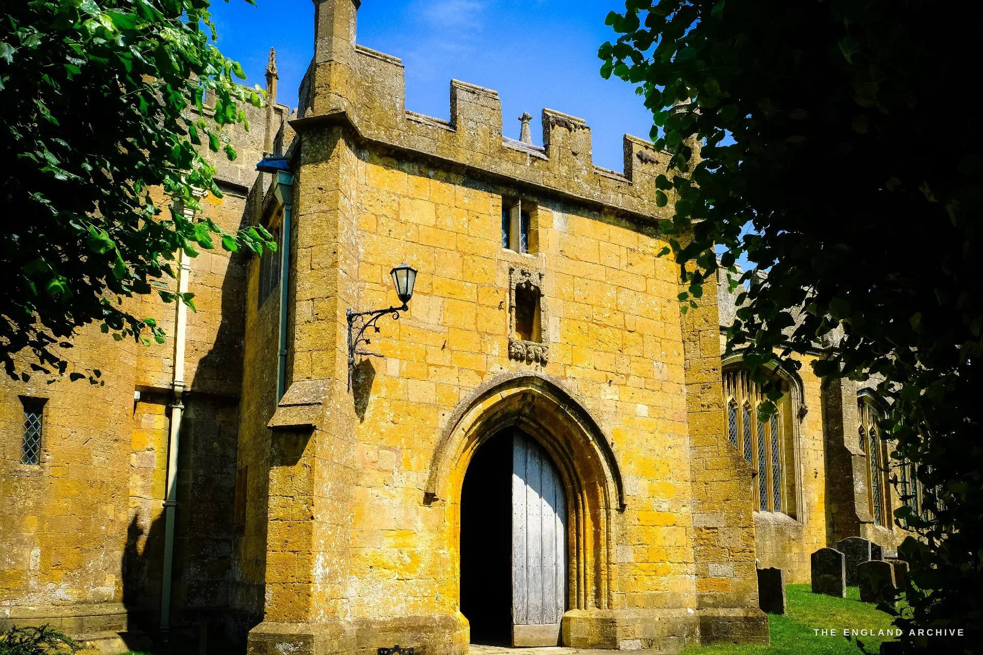 The battlemented honey-stone south porch of St James' Church, its pointed-arch door standing open, a wrought-iron lamp beside it and gravestones to the right.