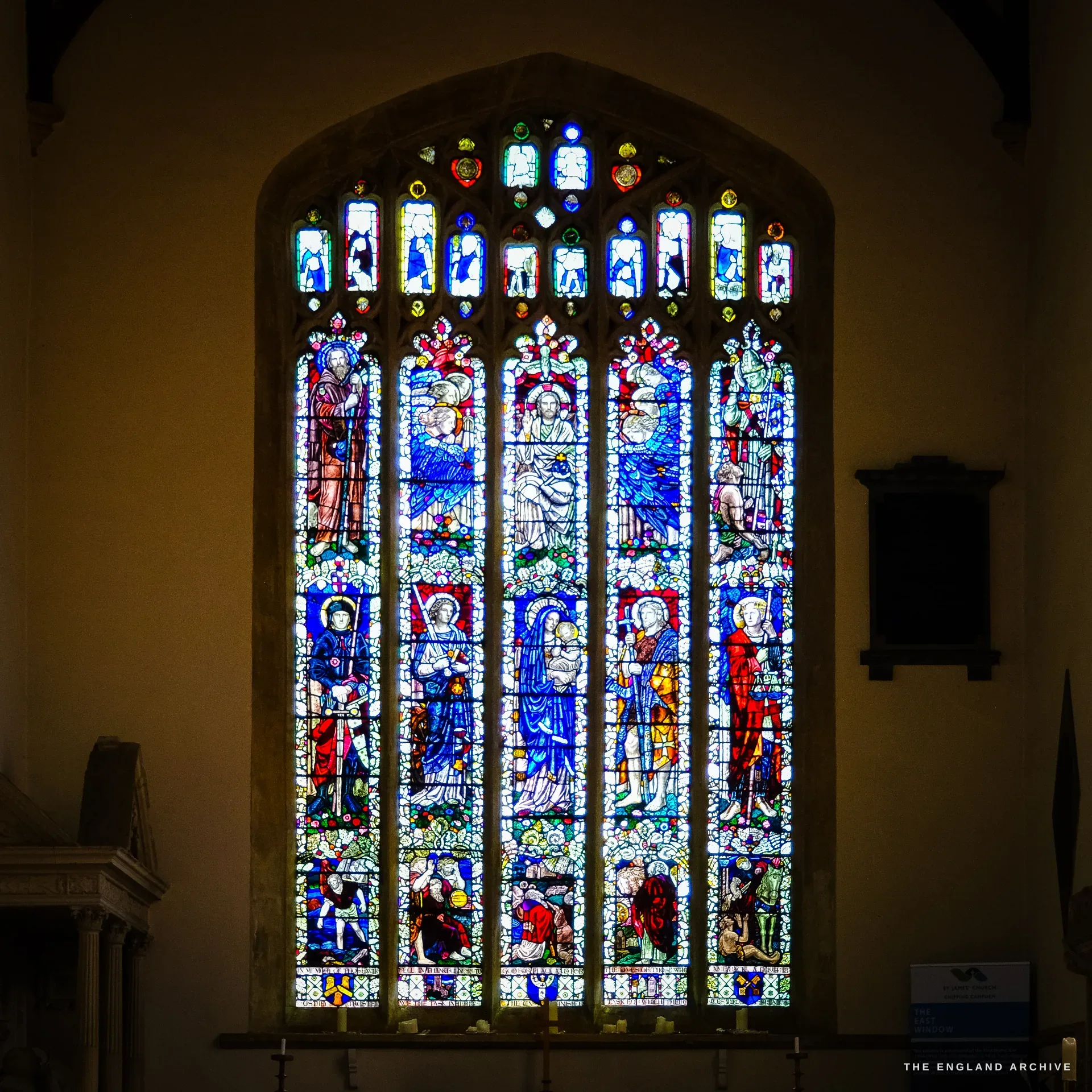 A tall five-light stained-glass window in St James' Church, two tiers of standing saints around a central Madonna and Child, candles lit on the sill below.