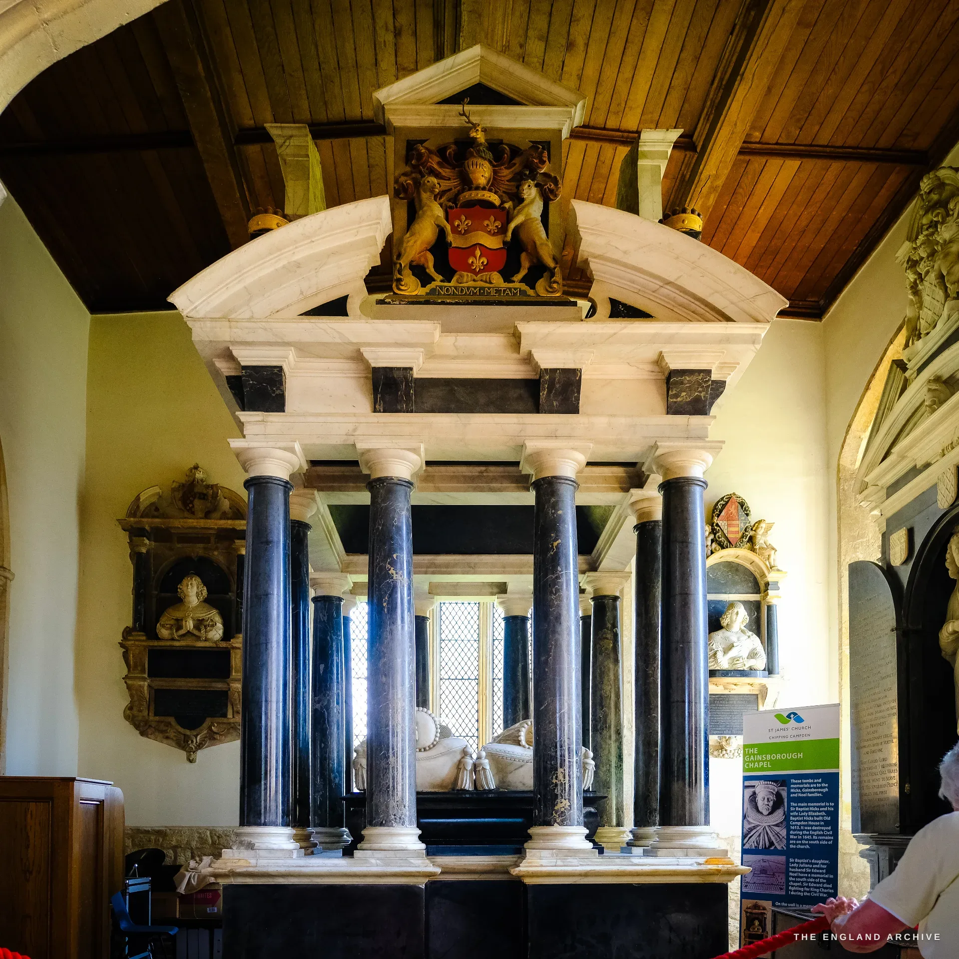 The great canopied table-tomb of Sir Baptist Hicks in the Gainsborough Chapel, black marble columns carrying a coffered canopy crowned with a painted achievement of arms, recumbent effigies beneath; Carol stands at the rope barrier to the right.