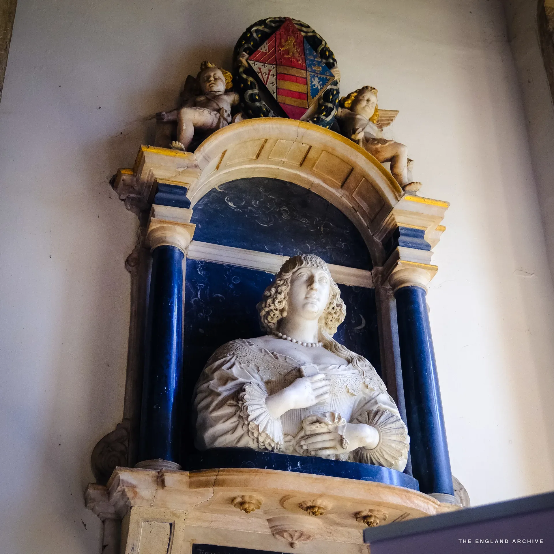 A white marble half-figure bust of a woman set in a deep blue niche flanked by blue columns, cherubs and a painted heraldic cartouche above.