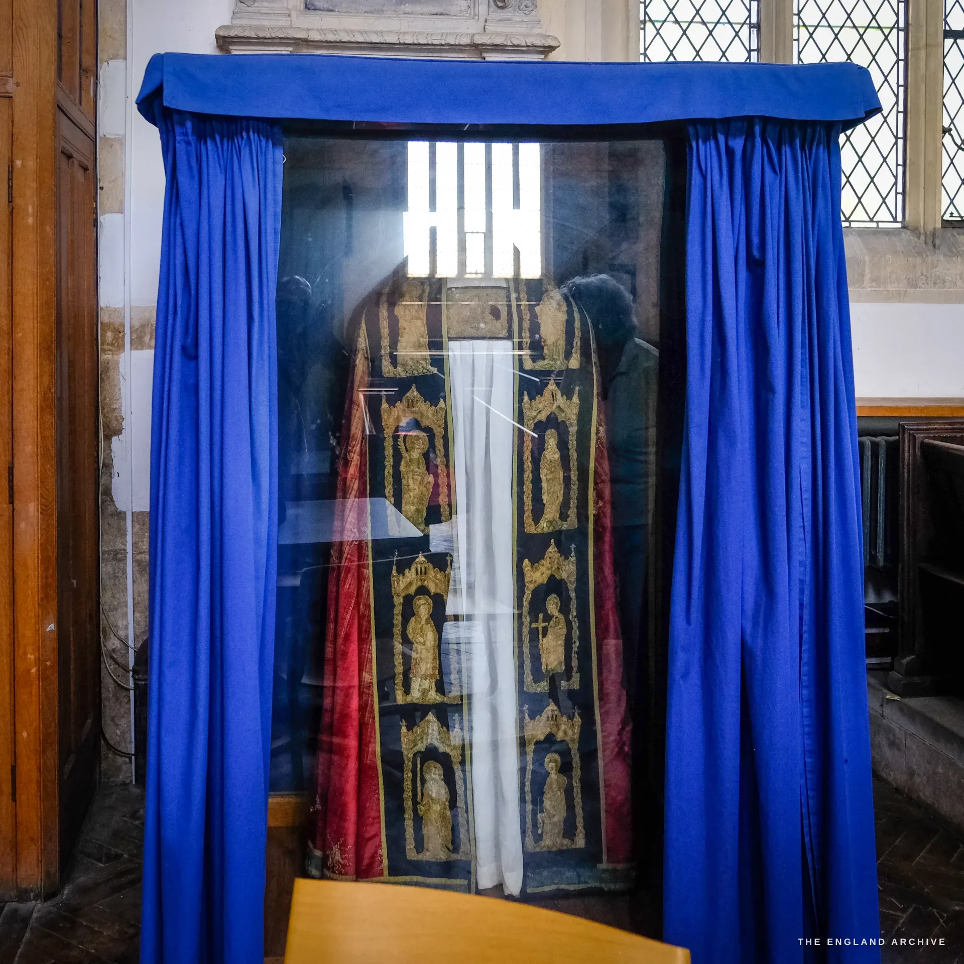 The glazed display case in St James' Church holding the medieval Campden altar hangings, gold-worked figures of saints on a dark ground behind reflective glass, blue curtains drawn back.