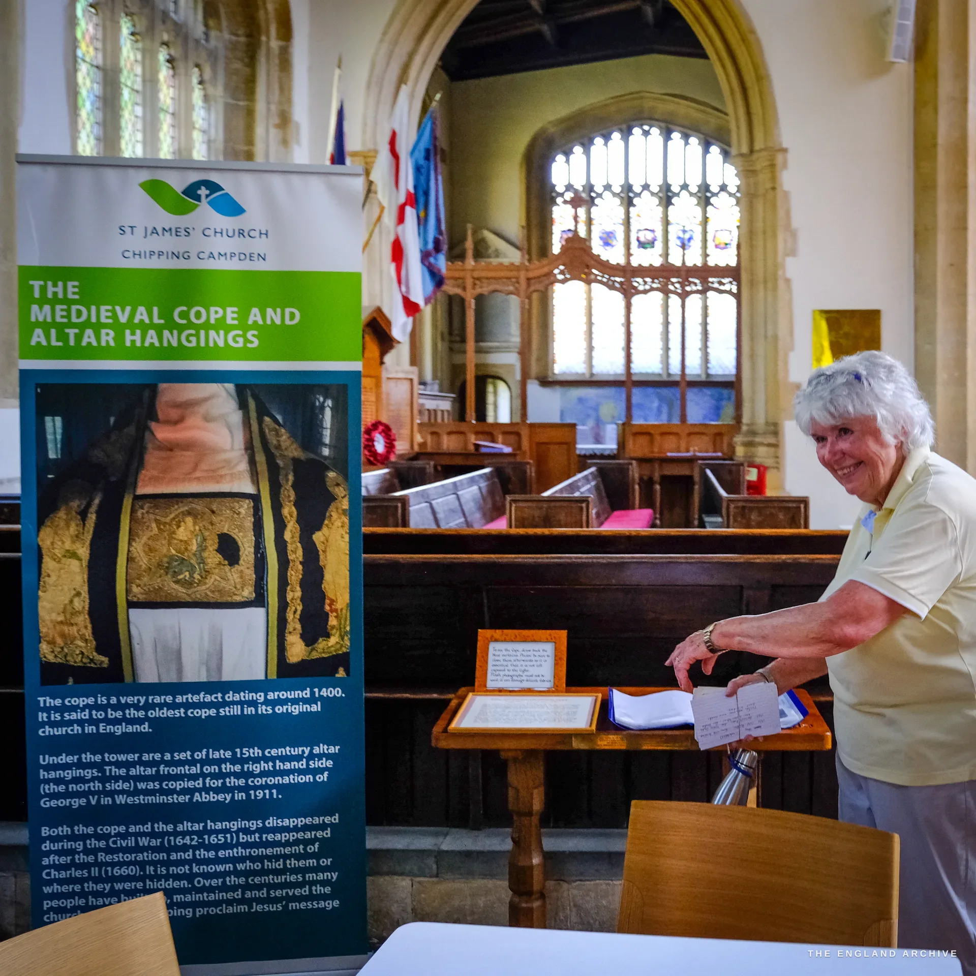 Carol stands smiling at the visitors' desk in St James' Church beside a tall banner about the medieval cope, pews and a stained-glass window behind her.