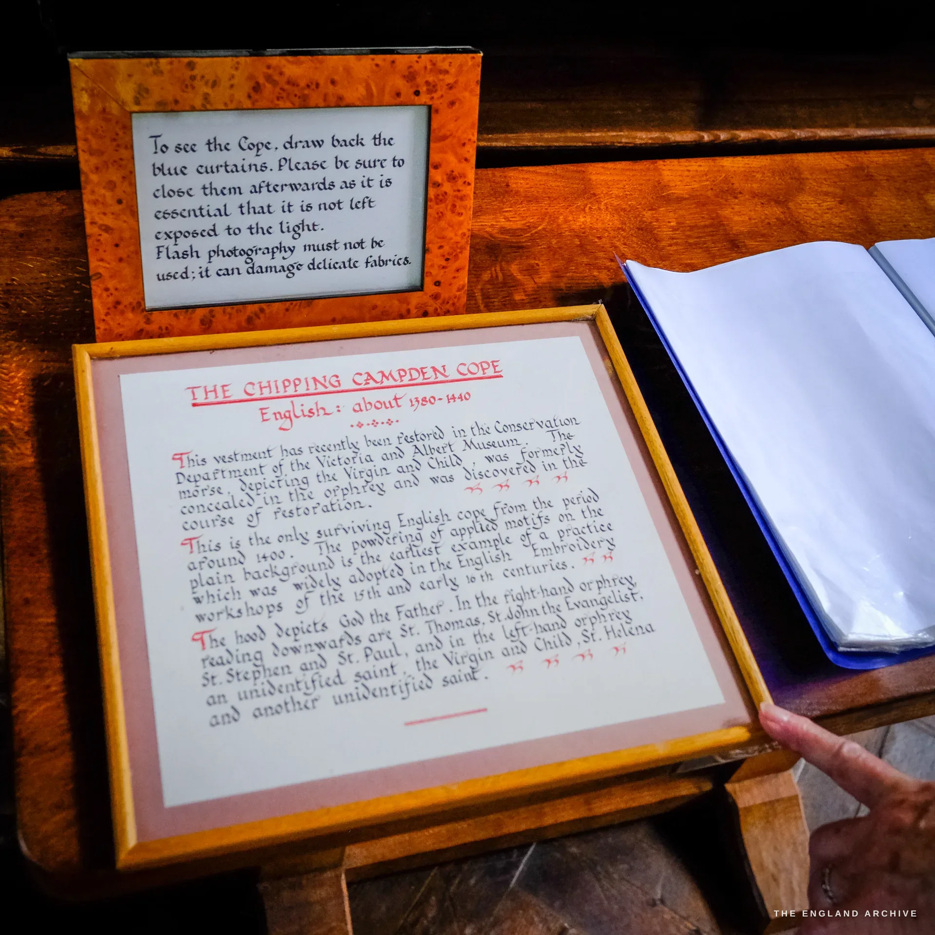 Two framed information cards and an open visitors' book on a wooden desk, one card asking visitors to draw back the blue curtains to see the cope and to use no flash, the other describing the Chipping Campden Cope of about 1380 to 1440.