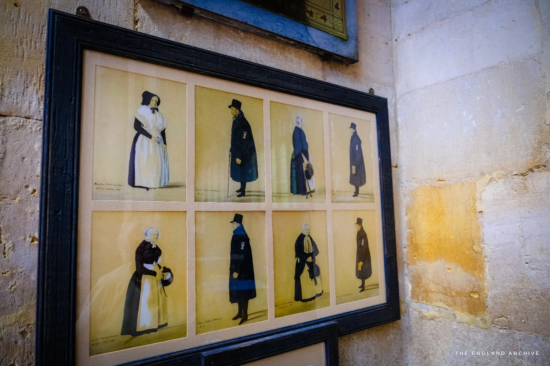 A black-framed set of eight small watercolours of the Campden almshouse occupants of the 1800s, in period dress, hung on a stone wall.
