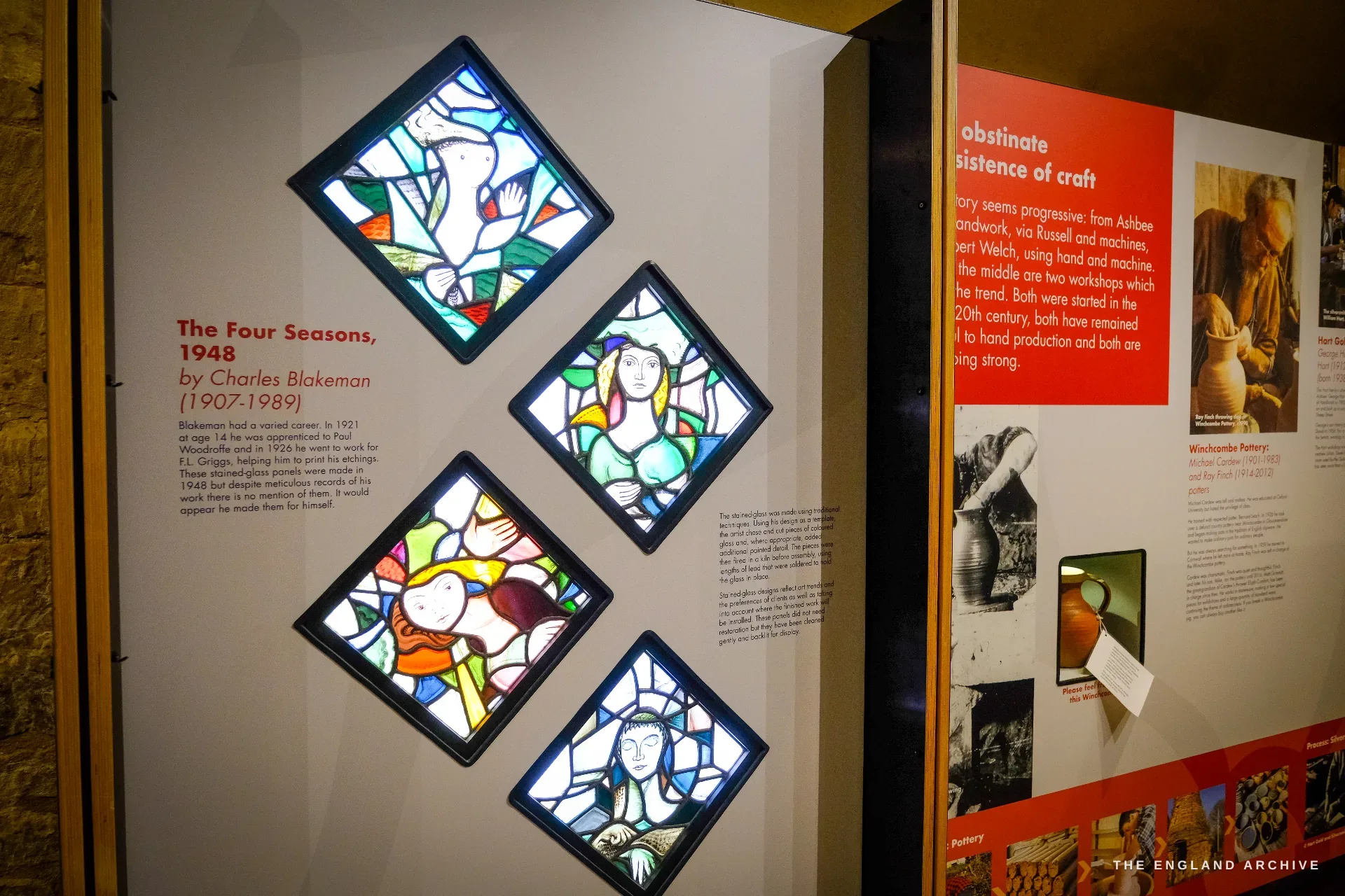 A museum display of four diamond-set stained-glass panels - Charles Blakeman's 'Four Seasons' of 1948 - mounted beside a red exhibition panel about Campden craft.