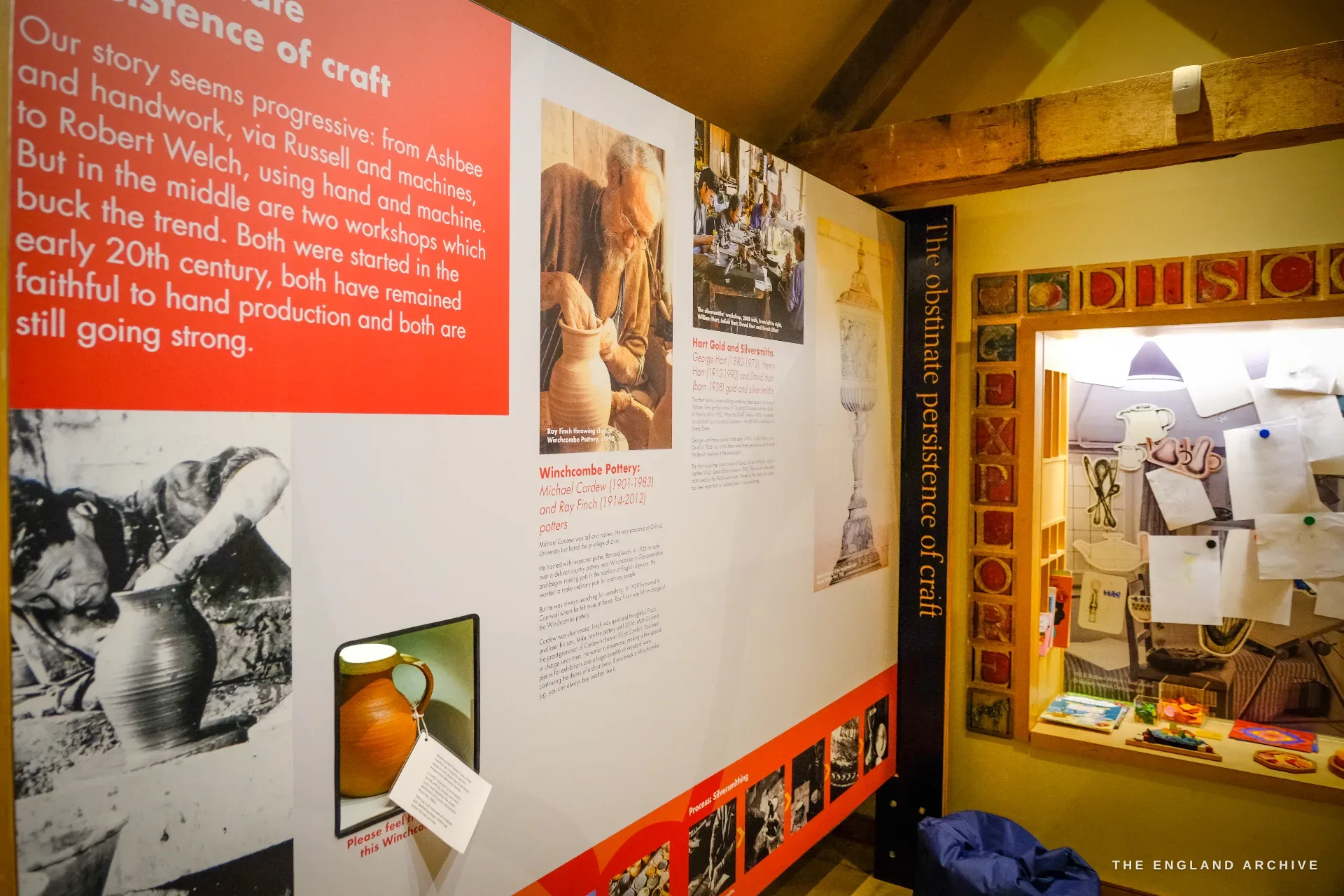 A wider view of the craft exhibition, a red panel headed 'The obstinate persistence of craft' carrying black-and-white photographs of a potter at the wheel beside a doorway marked 'Discover'.