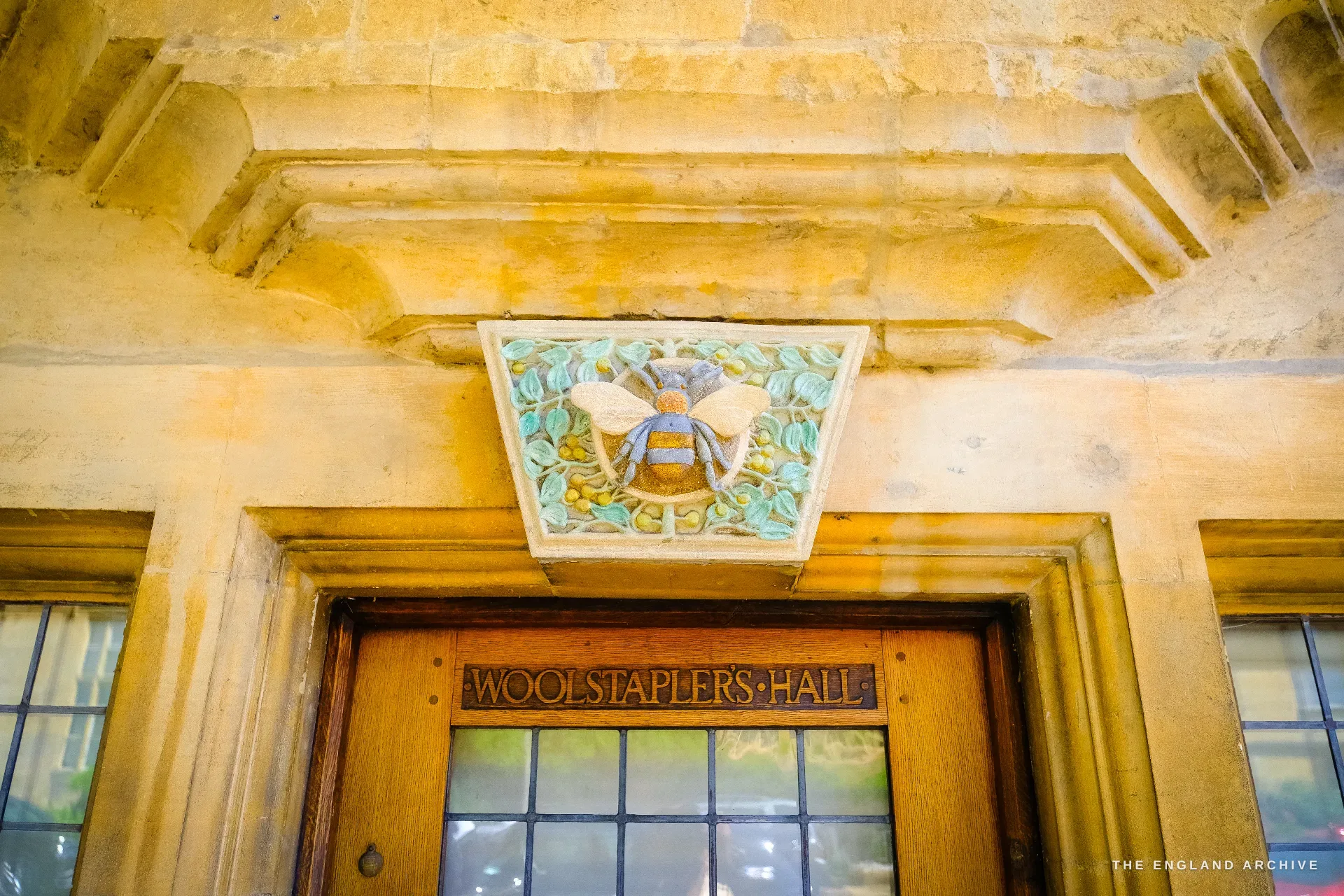 A carved and painted bee corbel over a doorway, the lintel below incised 'Woolstaplers Hall'.