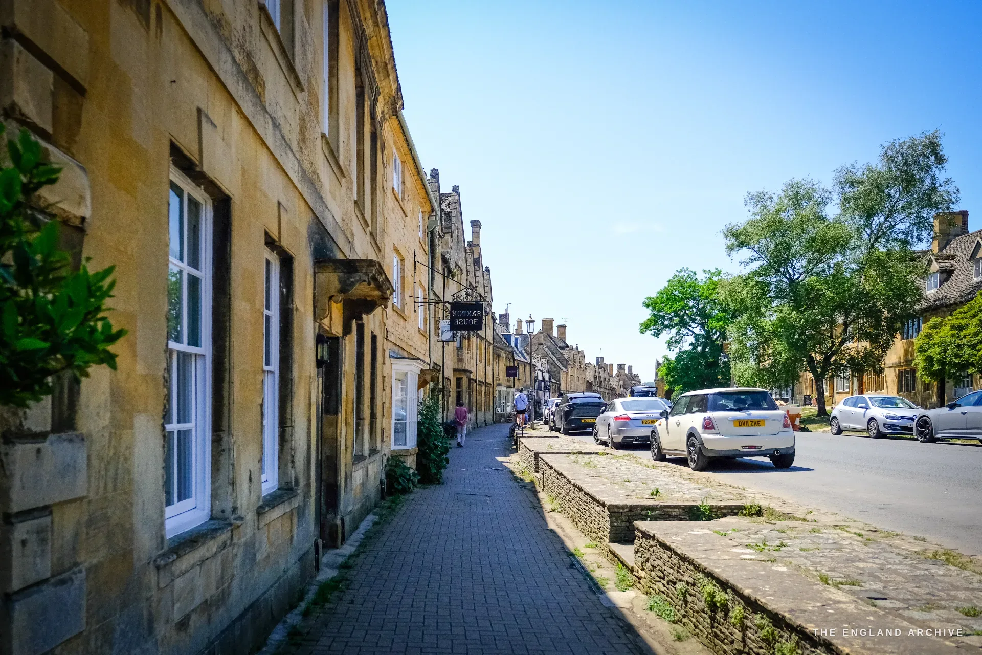 A classic High Street view, a terrace of honey-stone houses along a flagstone pavement behind a low stone wall, a hanging shop sign and parked cars under a tree-lined street.