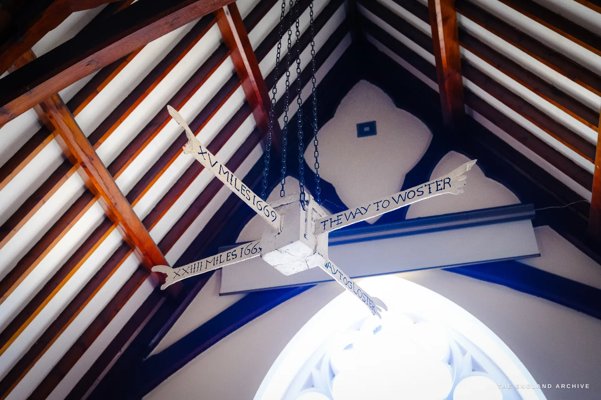 The same four-armed hanging direction sign seen from below against a peaked roof and a domed lantern, the arms dated 1669.
