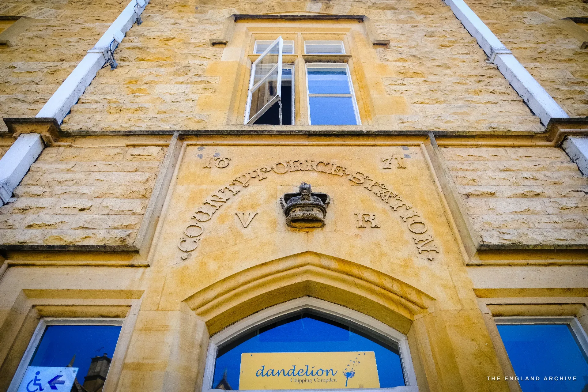 Looking up at a Victorian Gothic public building, a carved crown over the arched stone doorway incised 'County Police Station' and dated 1871 with the cypher 'V R' beside it.