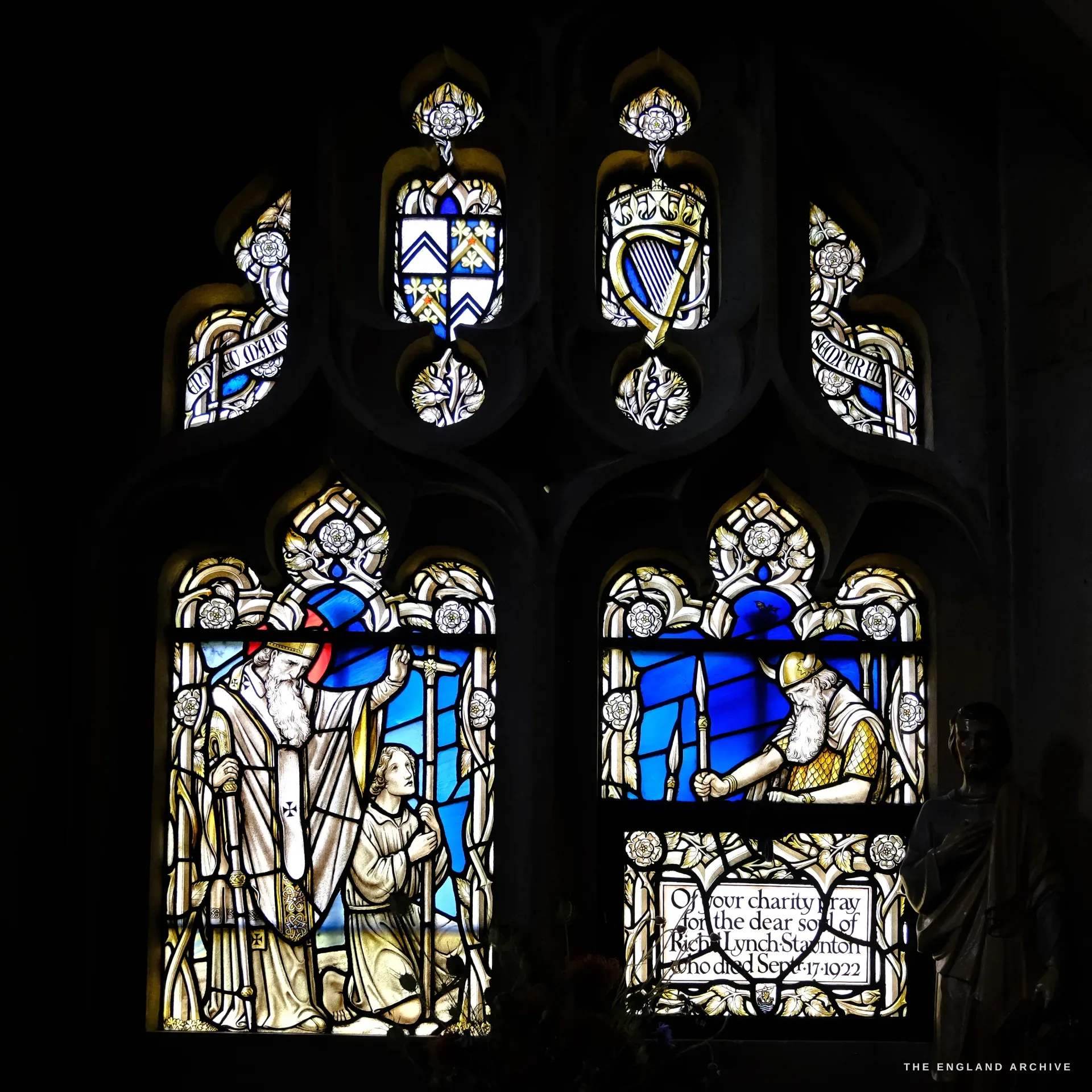 A two-light stained-glass window of bishops and saints with kneeling figures, a memorial panel below asking prayers for Richard Lynch Staunton, who died in September 1922.
