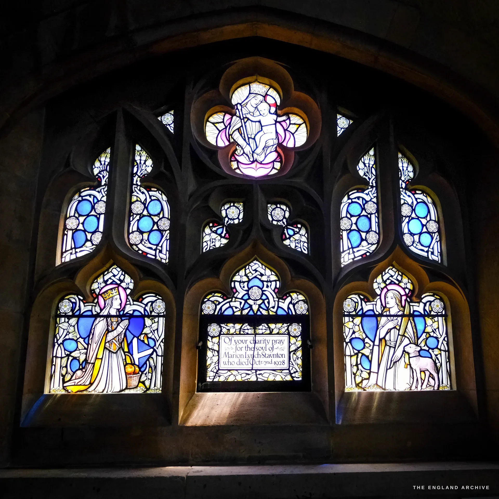 A three-light stained-glass window with a Madonna and Child in the tracery above, two crowned female saints flanking a memorial panel asking prayers for Marion Lynch Staunton, who died in October 1928.