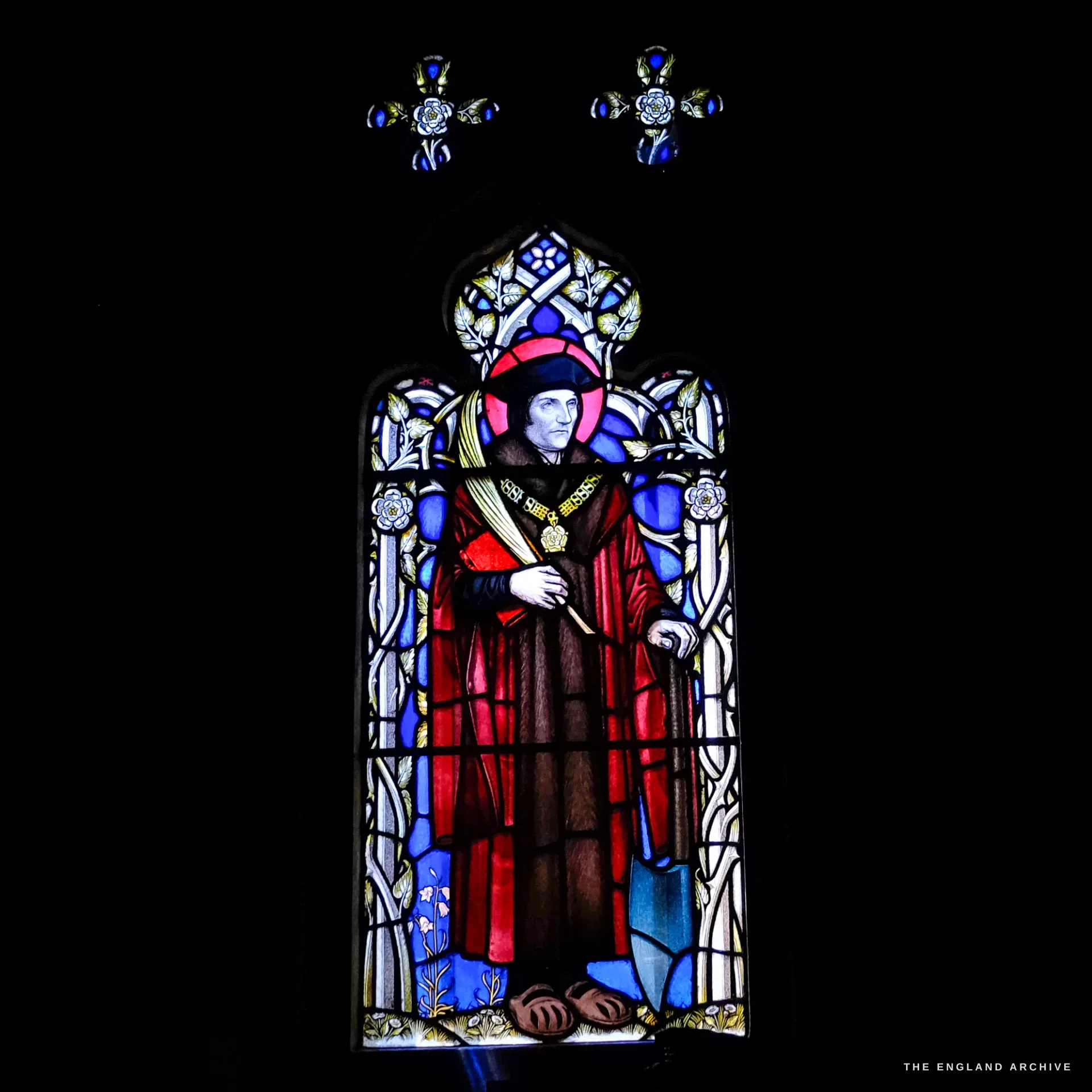 A single-light stained-glass window of a haloed saint in red and brown robes holding a martyr's palm and a spade, against blue and white floral quarries.