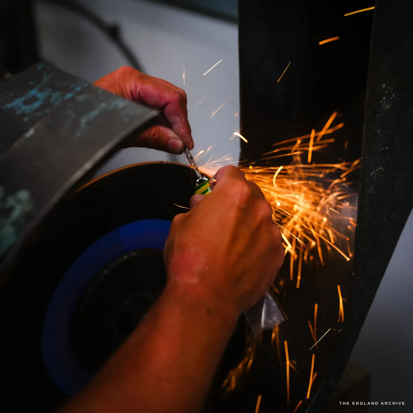 A close low-key frame of two hands holding a small piece to a fast-spinning wheel, a bright burst of orange sparks thrown off into the dark.