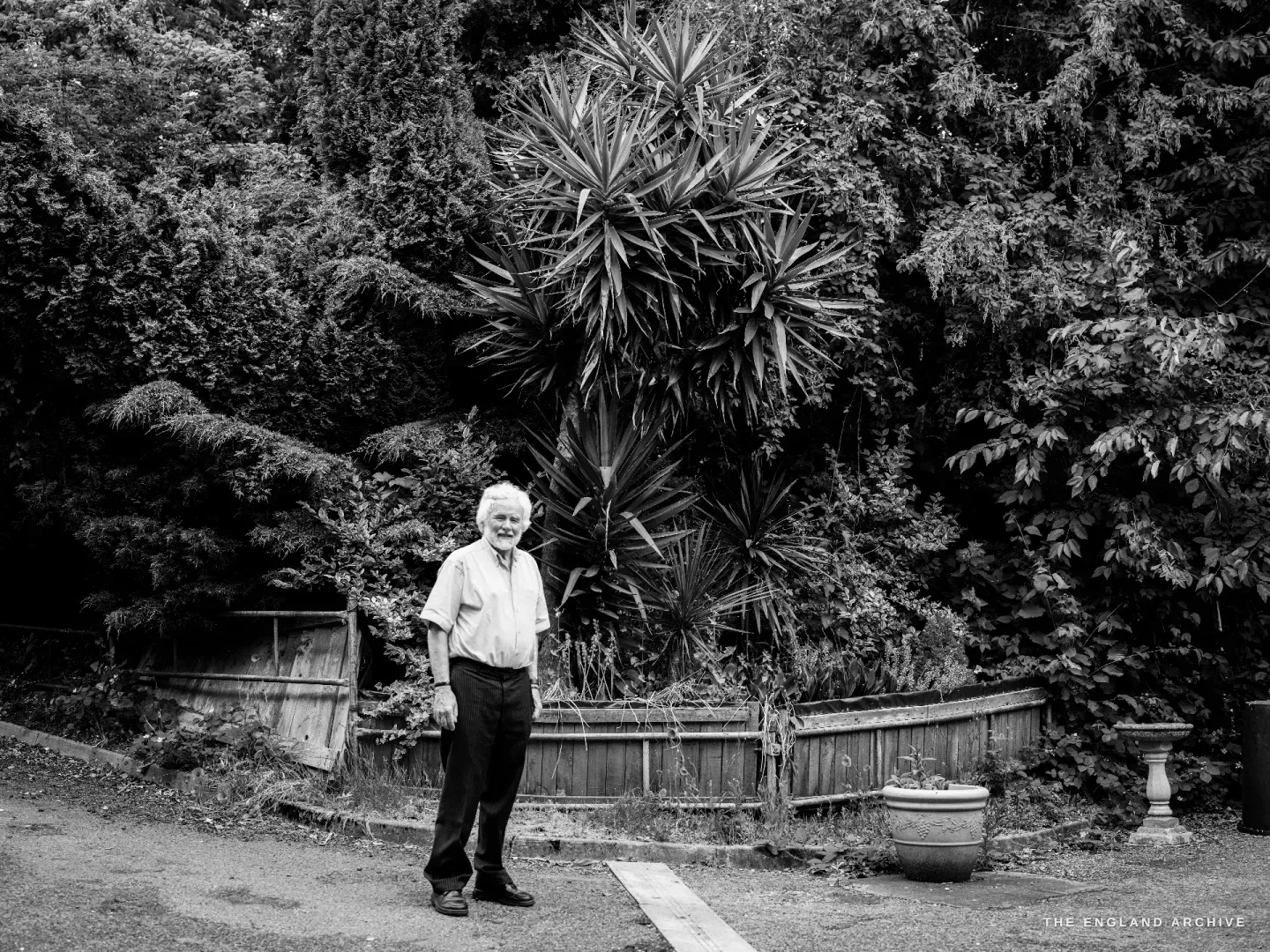 A full-length portrait of Chris Hudson standing in a green garden, white hair bright against the foliage, hands at his sides, a slight smile.