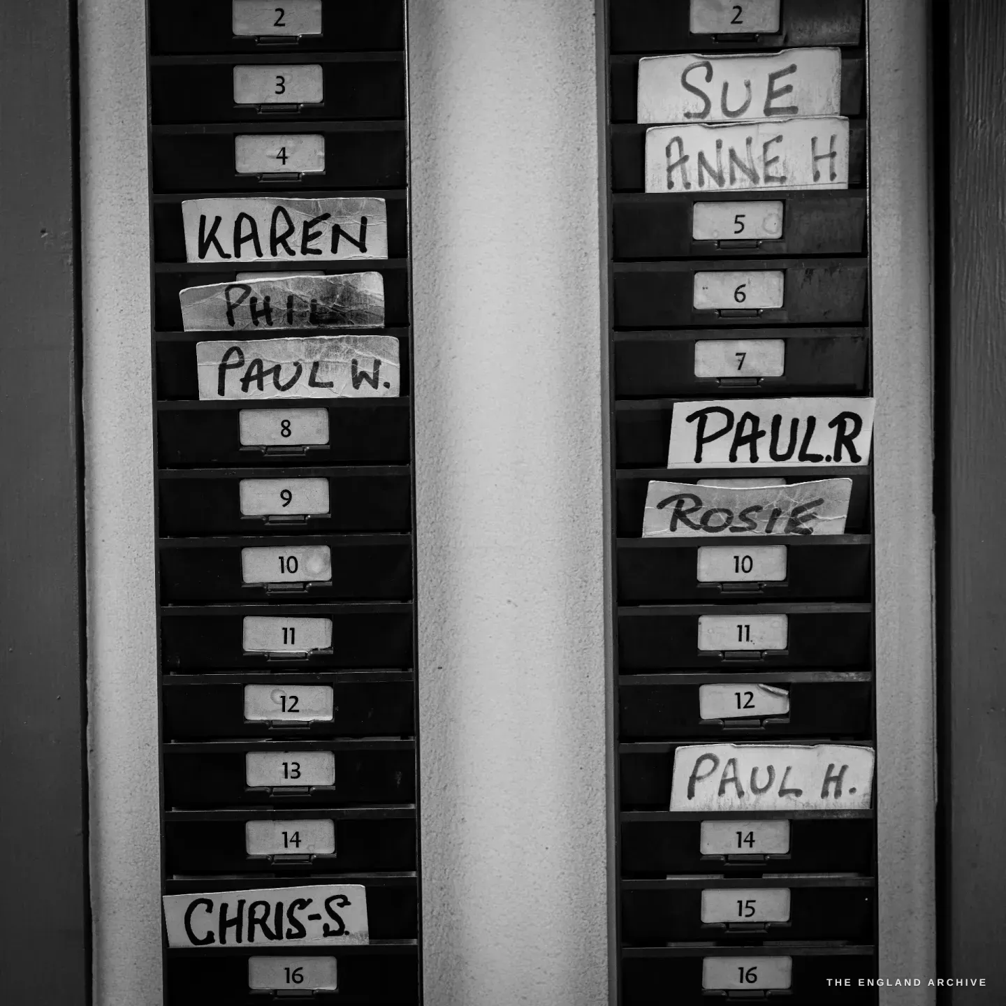 A clocking-in card rack on the wall, the slots hand-labelled with names - Karen, Phil, Paul W, Sue, Anne H, Paul R, Rosie, Paul H, Chris S.