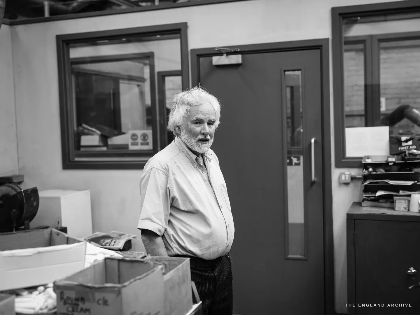 Chris Hudson standing among boxes of work on the factory floor, white hair and beard, looking to camera, a fire door and first-aid station behind him.