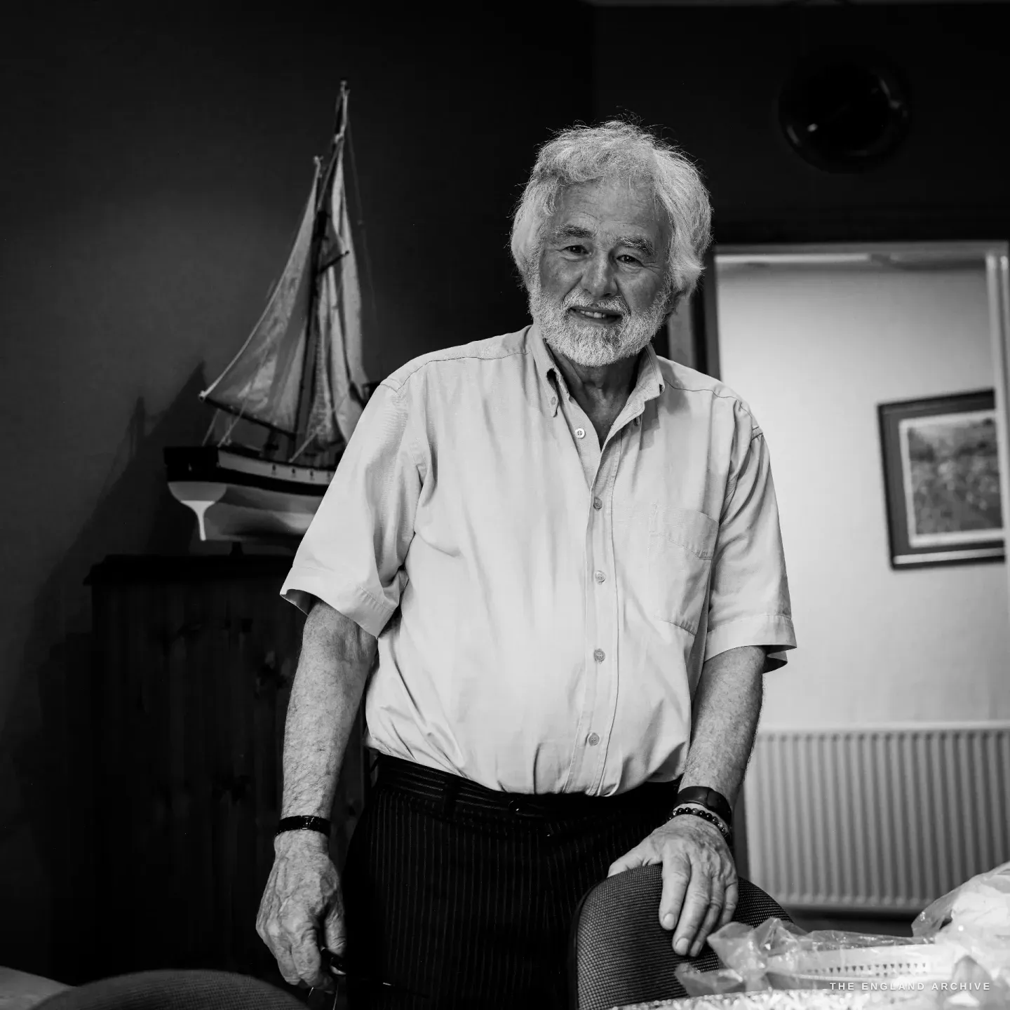 Chris Hudson standing in his office, white hair and beard, a warm smile, a model sailing ship on the shelf behind him.