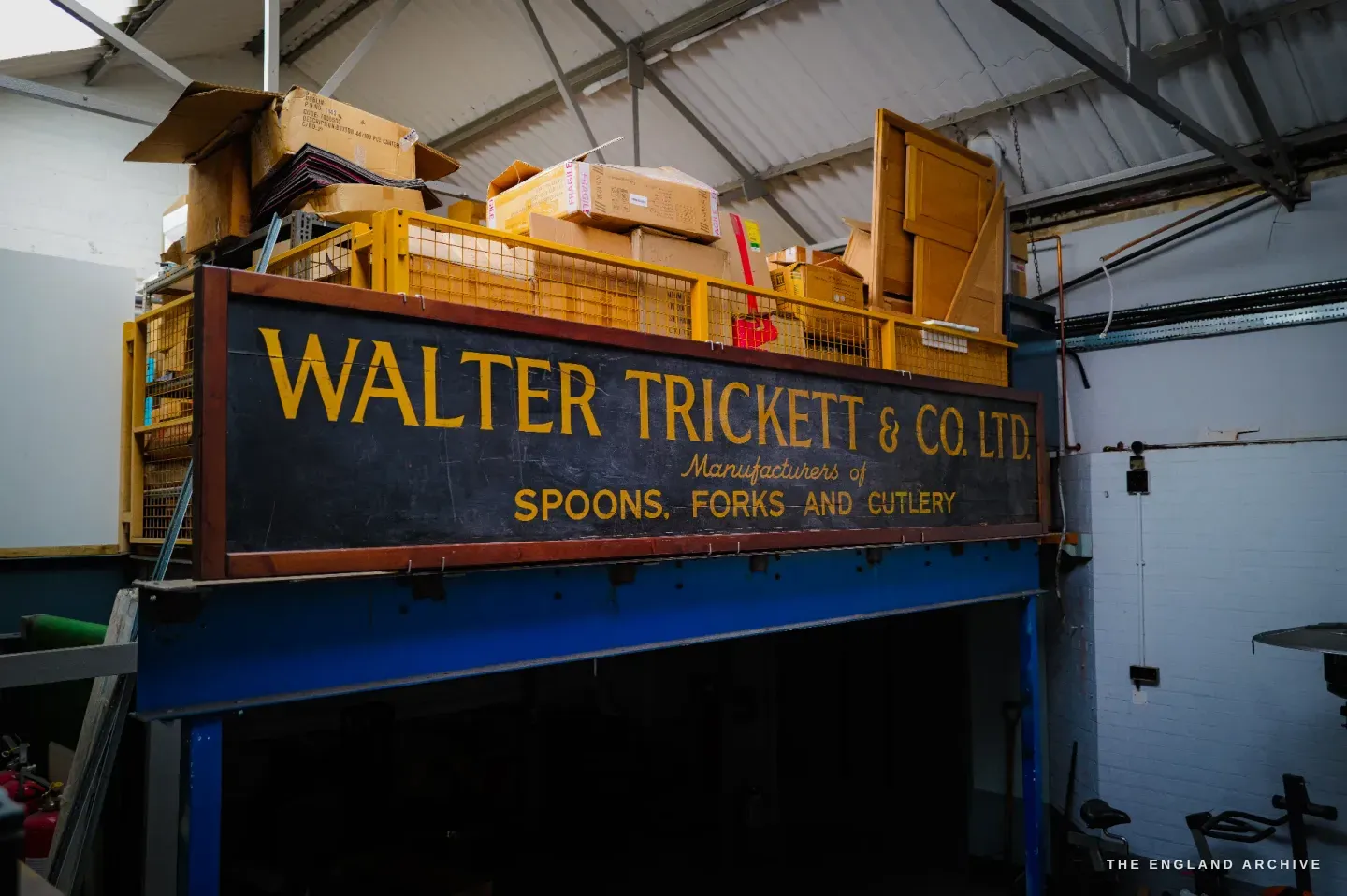 A large old painted sign reading WALTER TRICKETT & CO LTD, Manufacturers of Spoons, Forks and Cutlery, mounted high on the workshop wall above storage.
