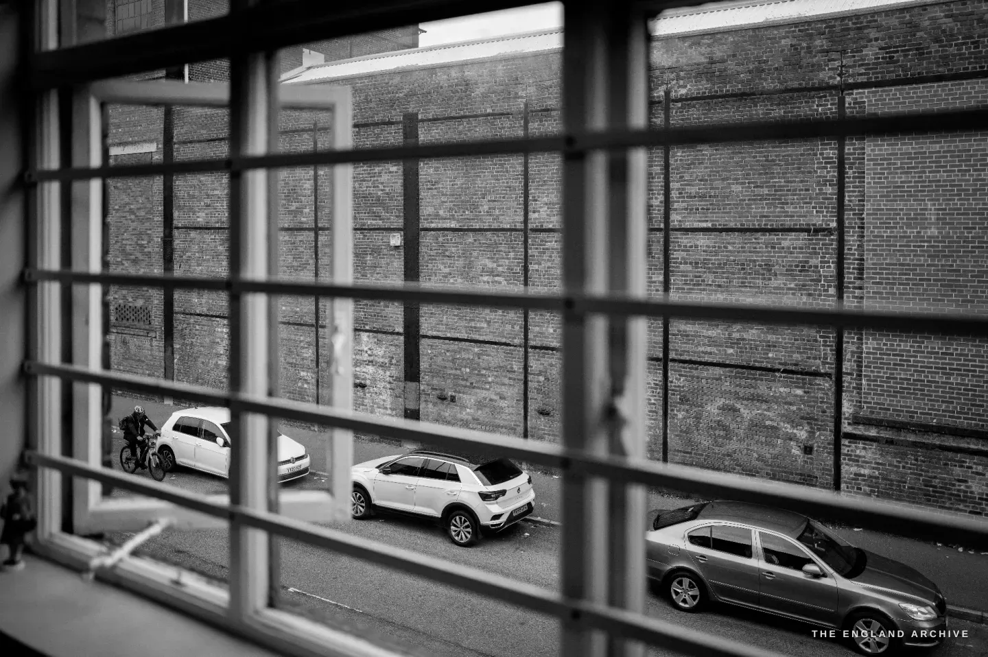 A view through a barred workshop window onto a Sheffield street: a long brick factory wall and parked cars below.