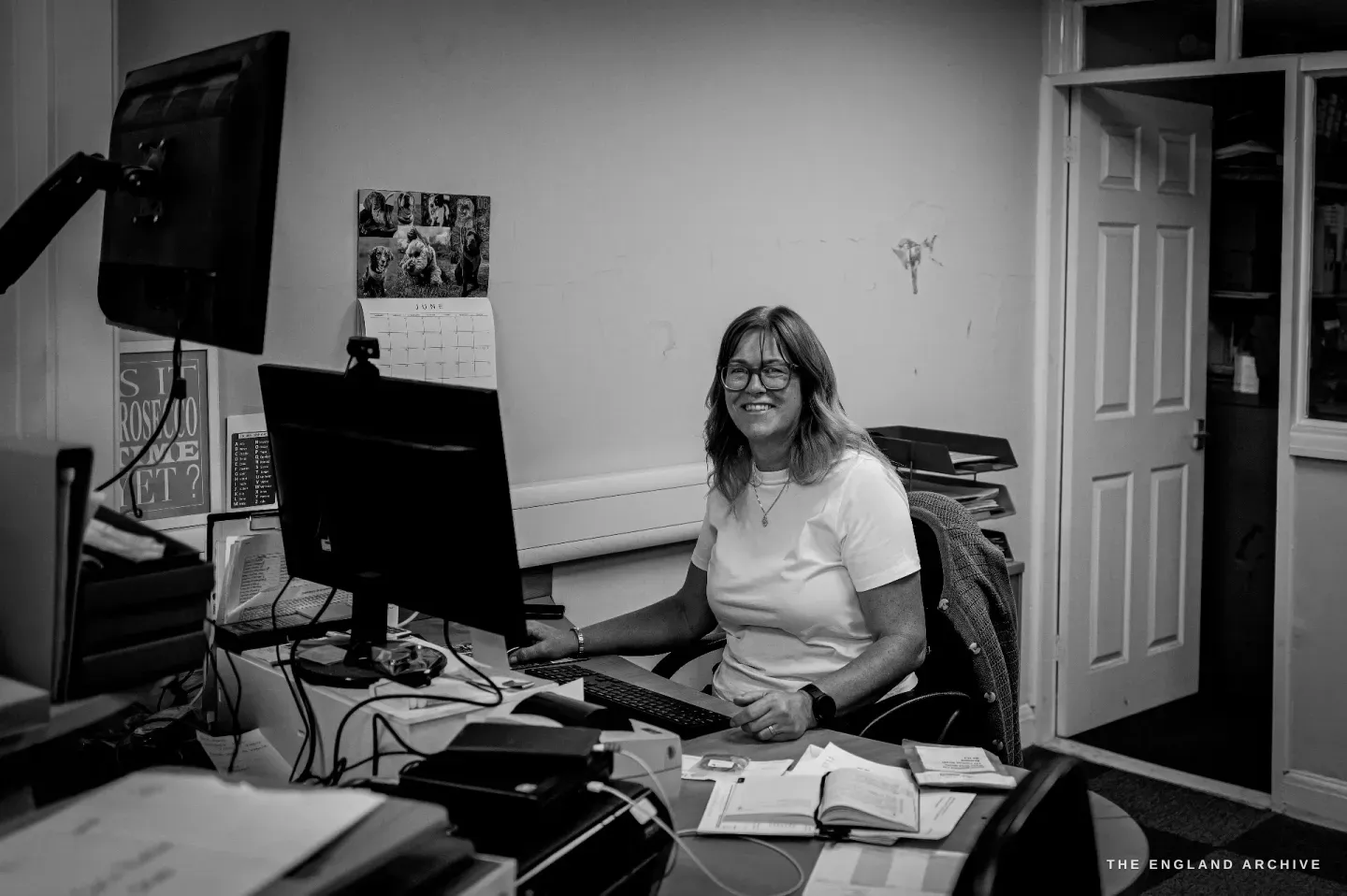 Karen seated at her office desk behind two monitors, smiling to camera, papers and ledgers spread in front of her.