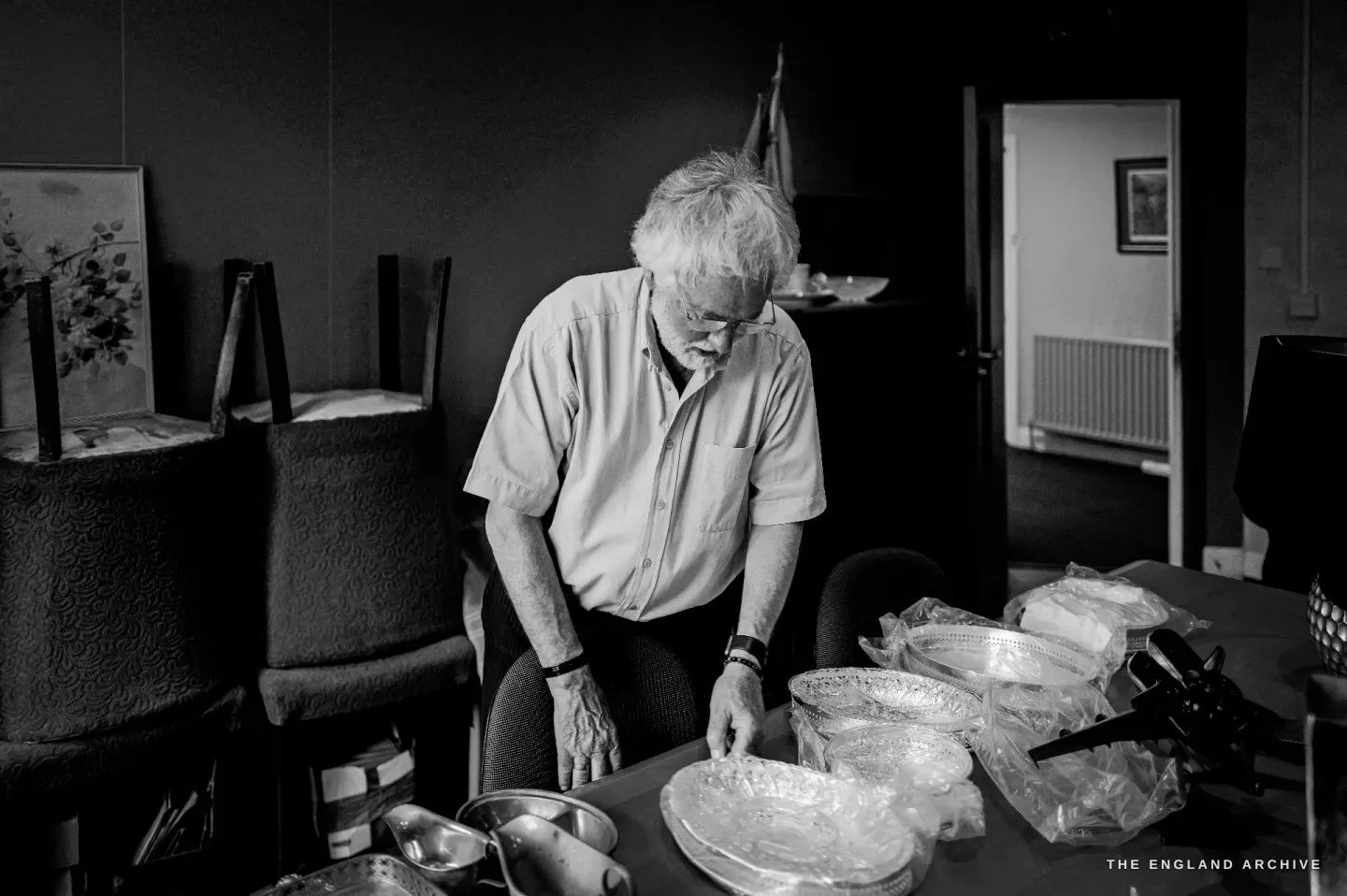 Chris Hudson leaning over a table laid with cut-glass and silver dishes, examining a piece, a framed picture and chairs stacked behind.