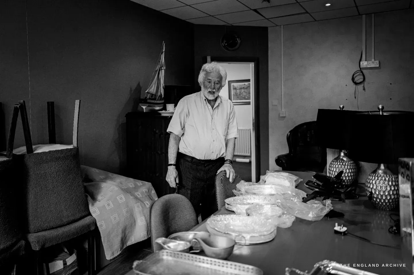 Chris Hudson standing at the head of a table set with silver and glass dishes, the model sailing ship and a framed landscape behind him.