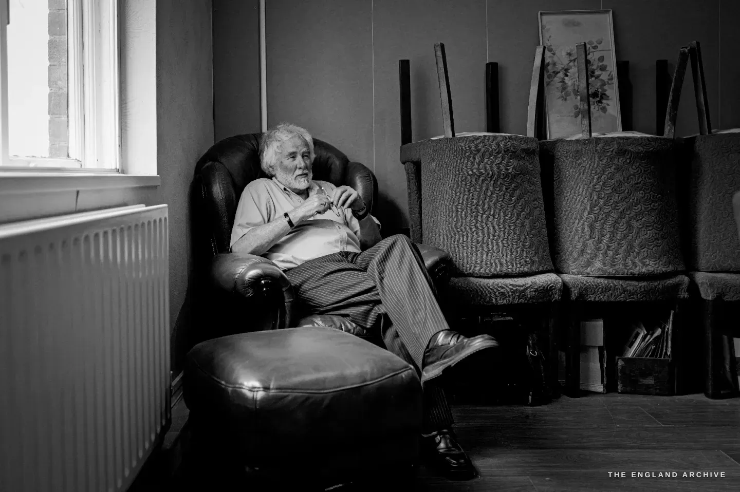 Chris Hudson sitting back in a leather armchair by a window, legs crossed on an ottoman, relaxed and smiling.