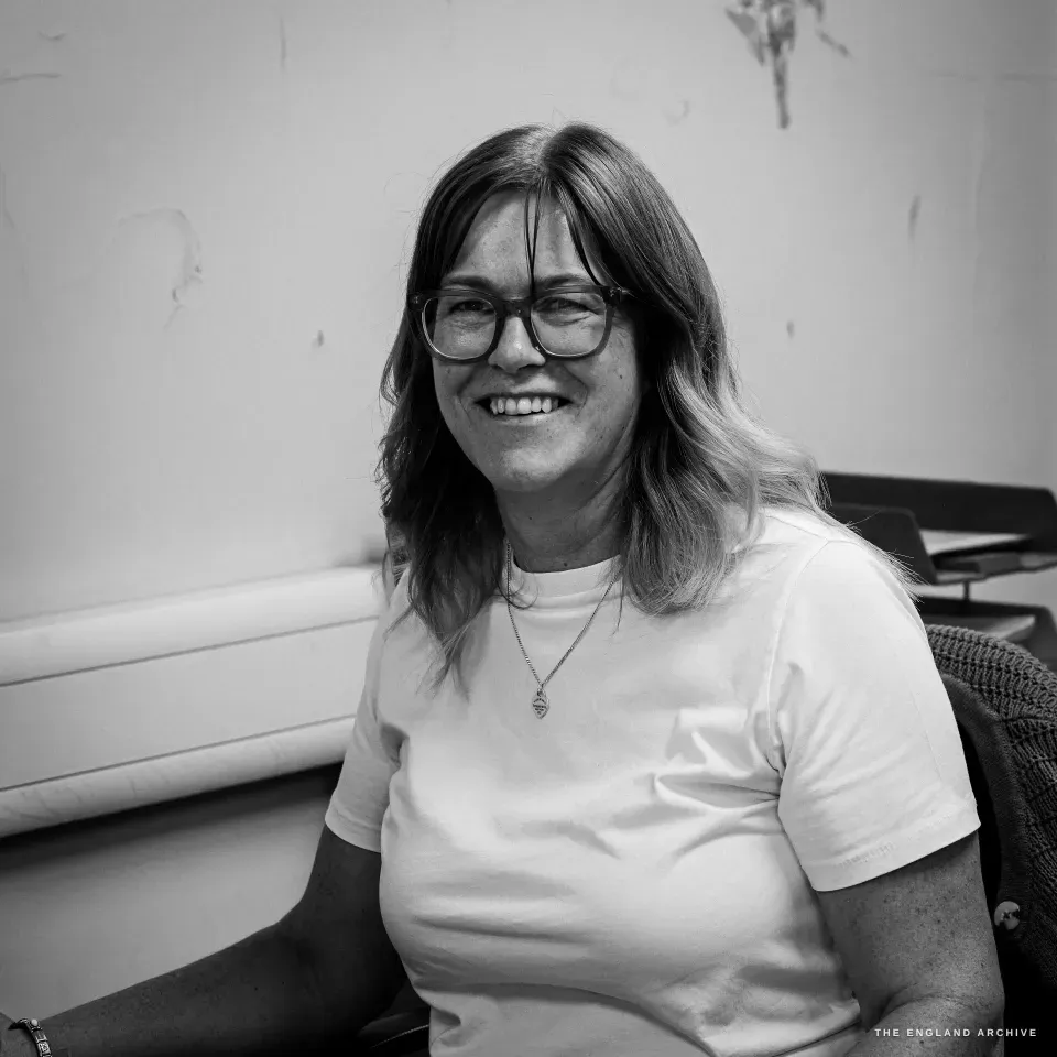 A close portrait of Karen, glasses and shoulder-length hair, smiling warmly to camera in the Chimo office.