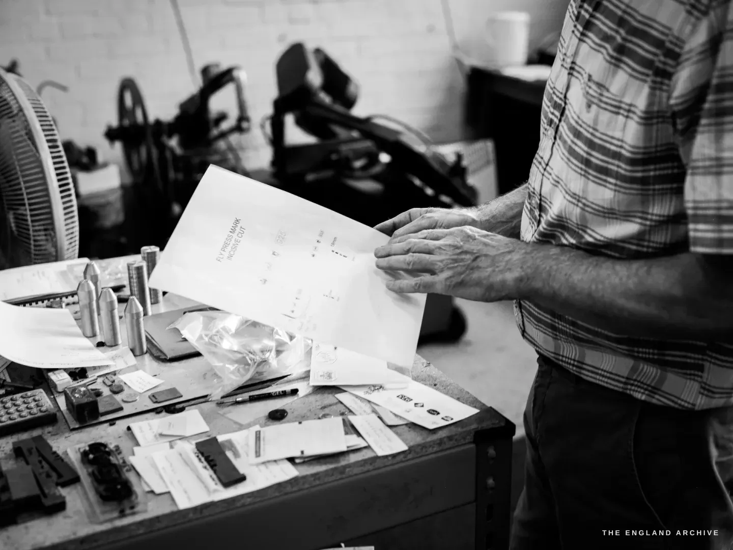 Chris Shaw at his bench holding a sheet of customer artwork, a tray of steel punches and small dies laid out in front of him.