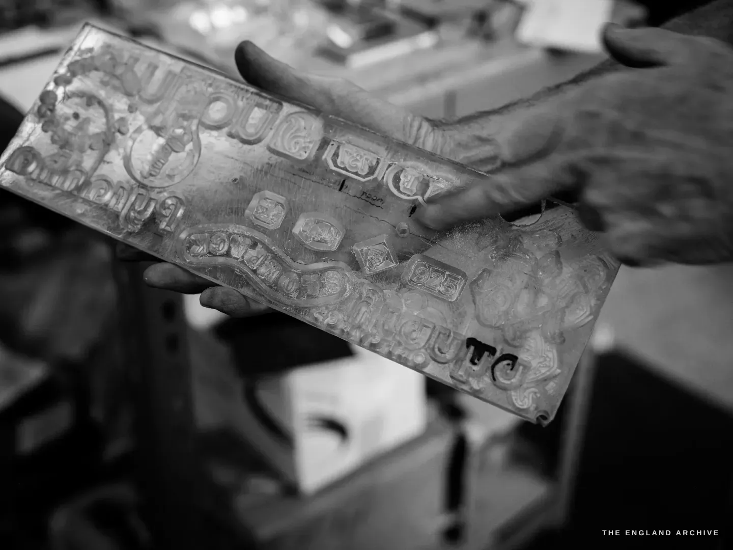 Two hands holding a long steel die plate cut with rows of letters, crests and ornamental panels in reverse relief.