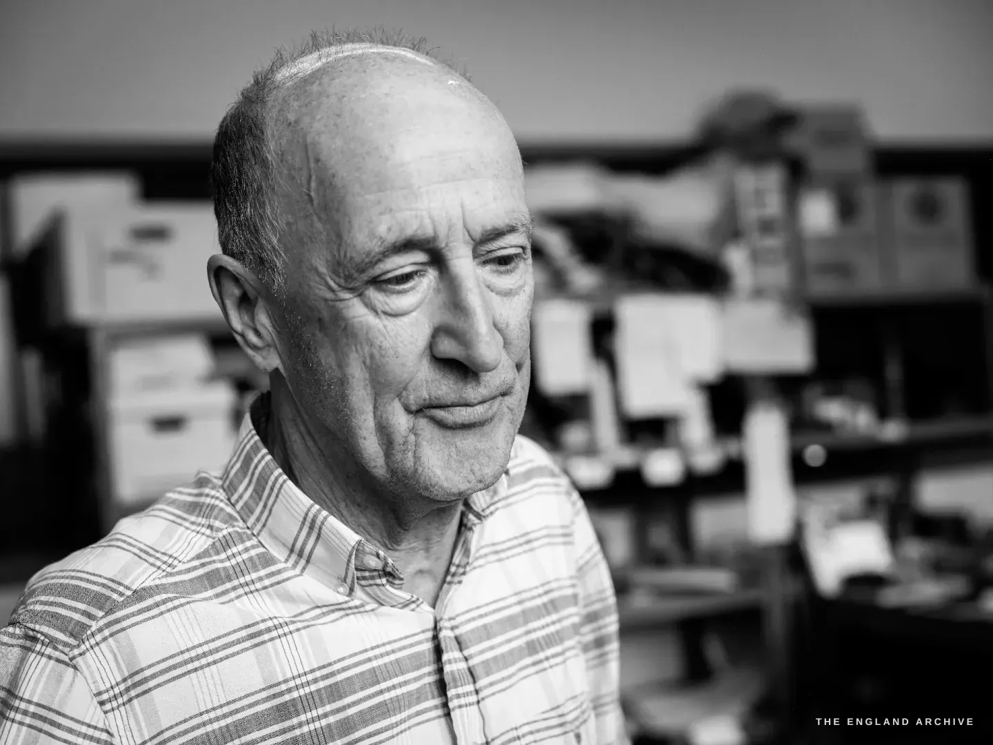 A close head portrait of Chris Shaw looking down in thought, the shelves of his workshop soft behind him.