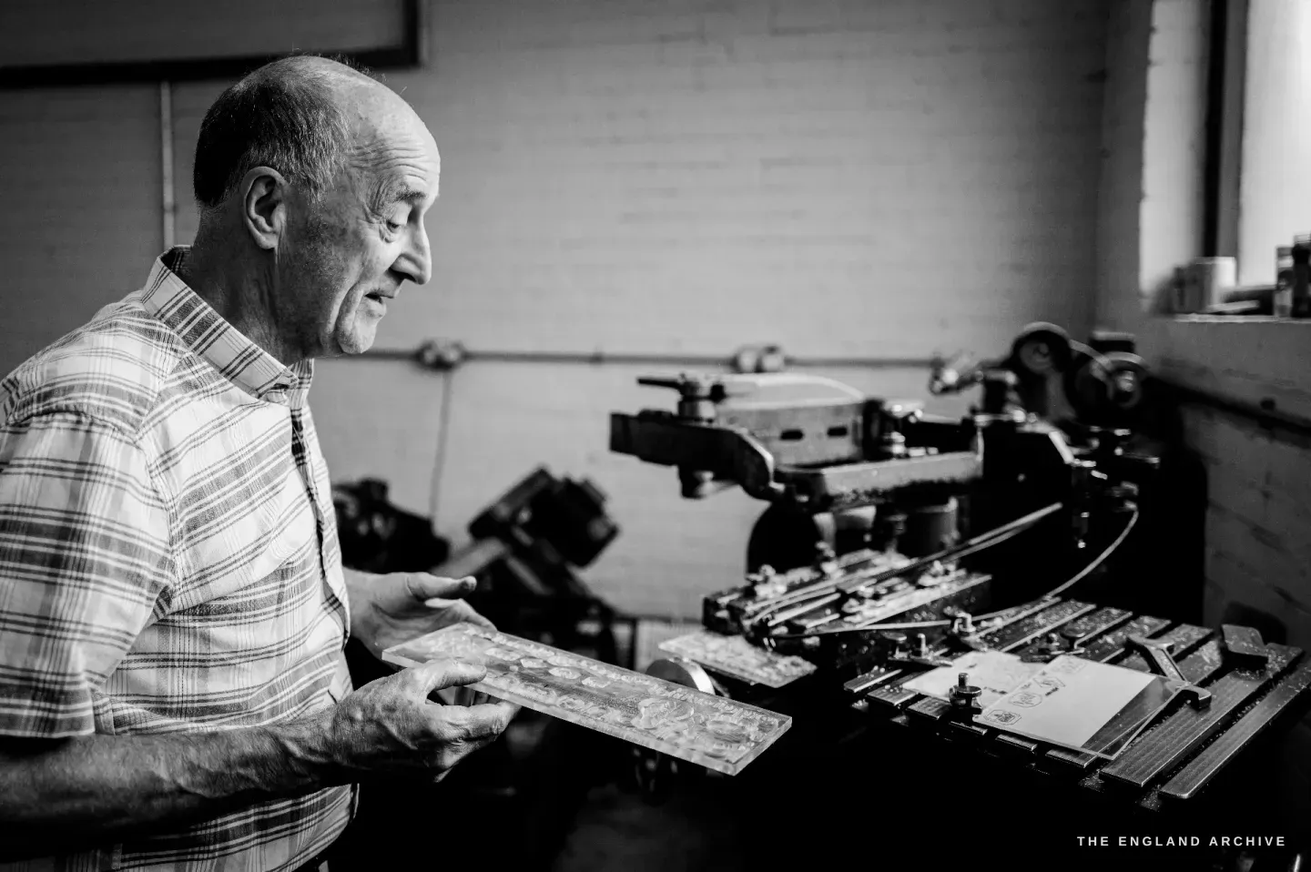 Chris in profile holding a steel die plate, studying it, the curved arm of a pantograph engraving machine on the bench beside him.