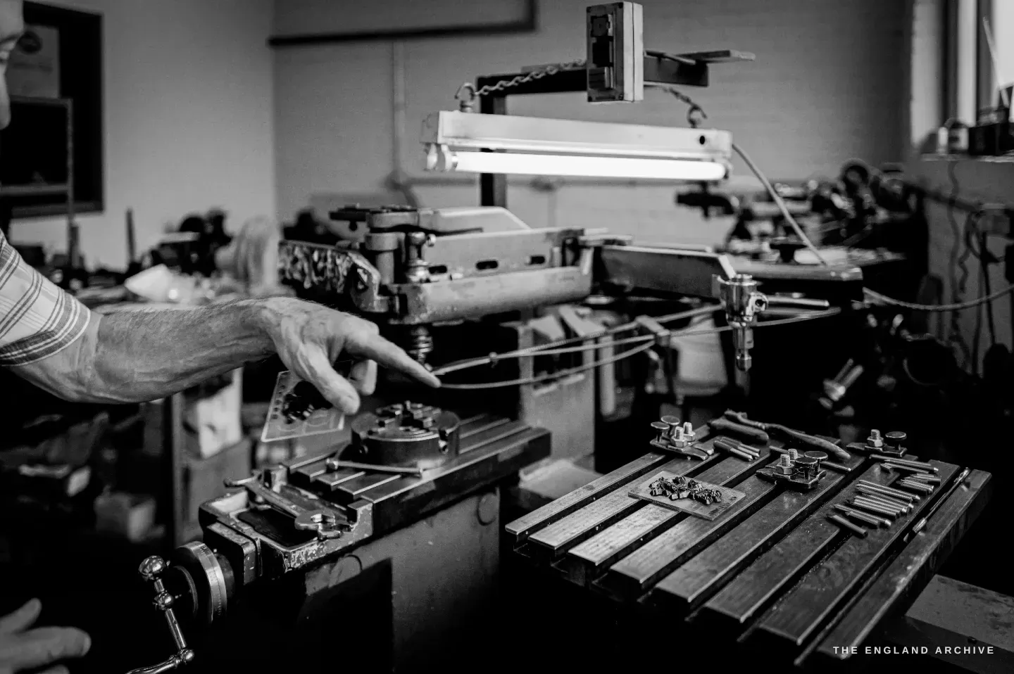 Chris’s hand at the controls of a pantograph engraving machine, the tracing arm and a circular work-holder lit under a work lamp.