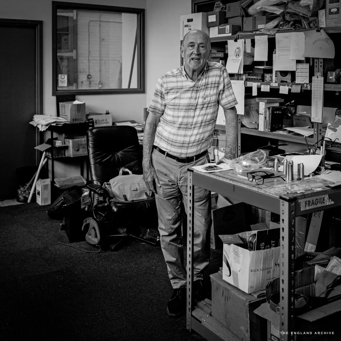 A full-length portrait of Chris Shaw standing easy beside his bench in the middle of the workshop, shelves of boxes and an office chair behind him.