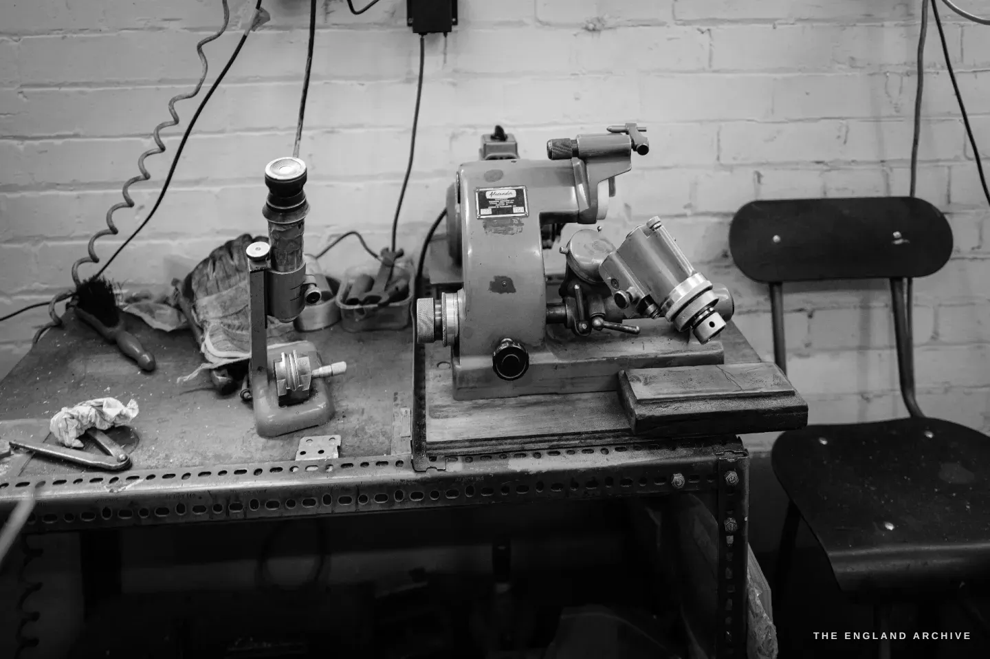 A pantograph engraving machine on its bench against a brick wall, its tracing arm, work-holder and microscope-like fittings catching the light.