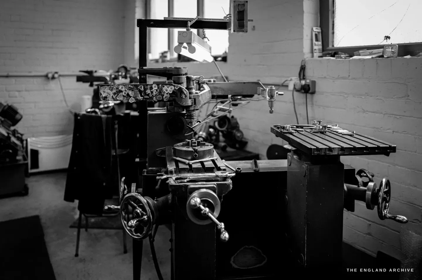 A pantograph engraving machine seen head-on under its work lamp, dies and fittings on the bed, the workshop receding behind.