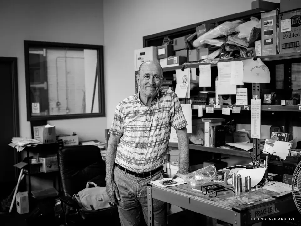 Chris Shaw standing at his workbench, leaning easily against it with a slight smile, shelves of boxes and tools behind him.