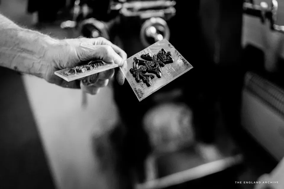 Chris holding up a small steel plate engraved with a heraldic beast in deep relief, the workshop dark behind.