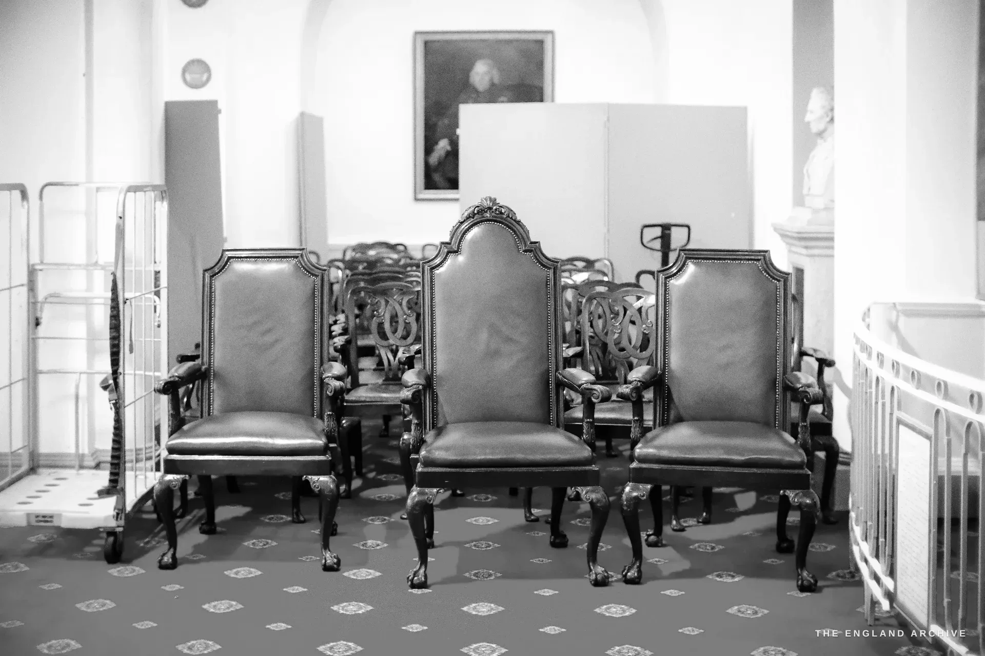 A group of red-leather ceremonial armchairs on a patterned red carpet, a portrait and a marble bust behind.